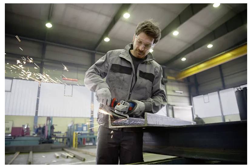 Ein Fabrikarbeiter in Schutzausrüstung benutzt ein Elektrowerkzeug, um ein Metallstück in einer Industriewerkstatt zu schleifen, wobei Funken fliegen.