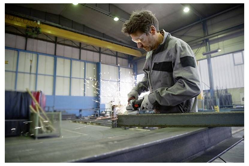 Eine Person in einer Werkstatt, die einen Schleifer auf einem Metallträger einsetzt, wobei Funken in einer großen Industrieanlage fliegen.