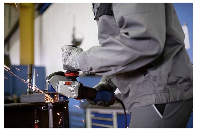Eine Person in Schutzkleidung und Handschuhen bedient einen Winkelschleifer an Metall und erzeugt dabei Funken in einer industriellen Werkstattumgebung.