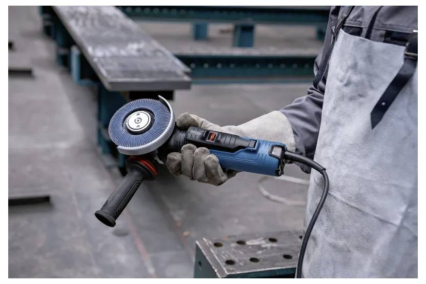 Eine Person mit Handschuhen und Schürze hält einen blauen Winkelschleifer und steht auf einem industriellen Boden mit Metallträgern im Hintergrund.