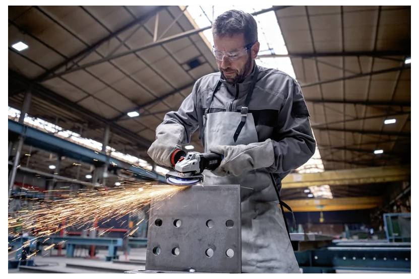 Ein Mann in Schutzausrüstung schleift ein Metallstück mit einem Elektrowerkzeug in einer Industriewerkstatt, während Funken sprühen und Maschinen im Hintergrund zu sehen sind.