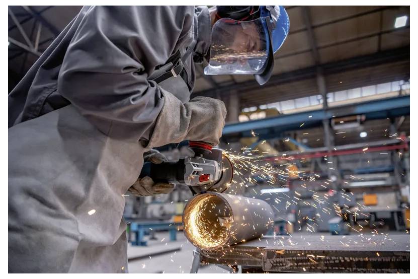 Ein Arbeiter mit Schutzausrüstung verwendet einen Winkelschleifer, um ein Metallrohr zu schneiden, wobei in einer industriellen Werkstattumgebung Funken entstehen.