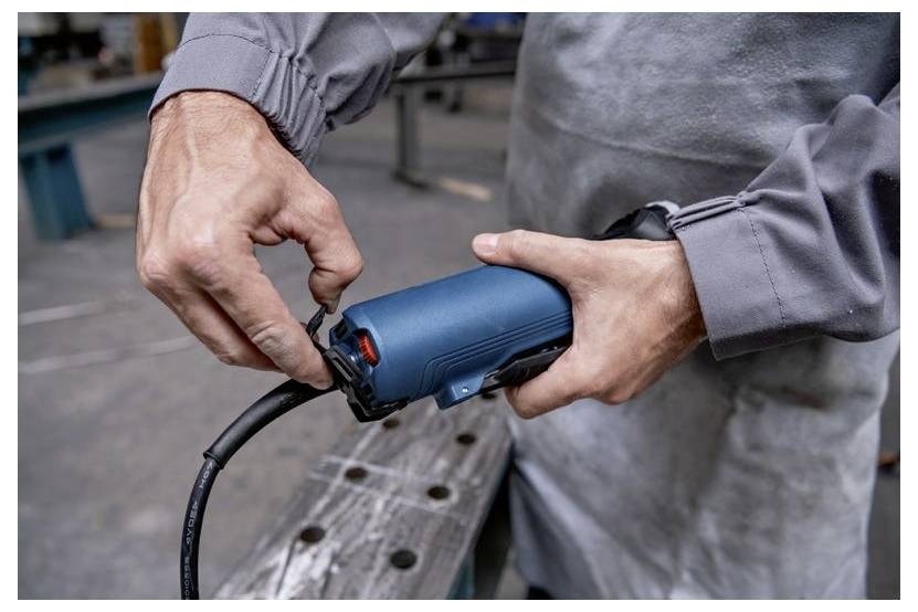 Eine Person in Arbeitskleidung benutzt ein blaues Elektrowerkzeug in einer Werkstatt und konzentriert sich darauf, Einstellungen vorzunehmen oder eine Komponente anzubringen.