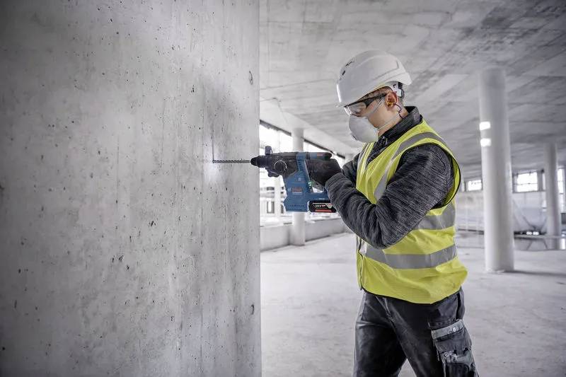 Ein Bauarbeiter mit Schutzhelm und Sicherheitsweste bedient eine Bohrmaschine an einer Betonwand im Innenraum eines unfertig gebauten Gebäudes.