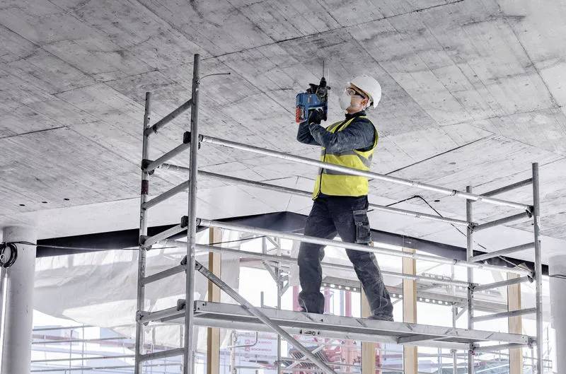 Ein Bauarbeiter in Sicherheitsausrüstung steht auf Gerüsten und bohrt in eine Betondecke auf einer Baustelle.