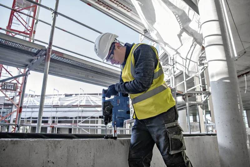 Ein Bauarbeiter mit gelber Warnweste und Schutzhelm bedient ein Elektrowerkzeug auf einer Baustelle mit Gerüsten und einem Gebäuderahmen.