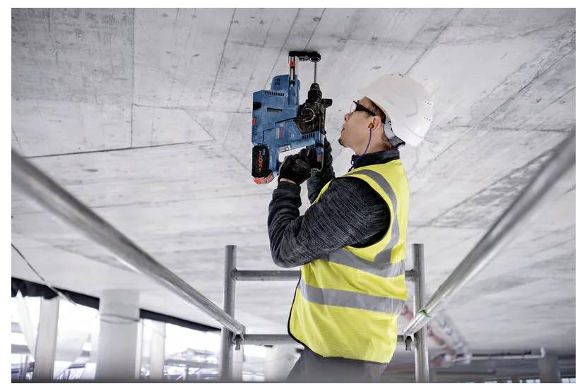 Ein Bauarbeiter in einer Sicherheitsweste und einem Schutzhelm verwendet ein Elektrowerkzeug, um in eine Betondecke auf einem Gerüst zu bohren.