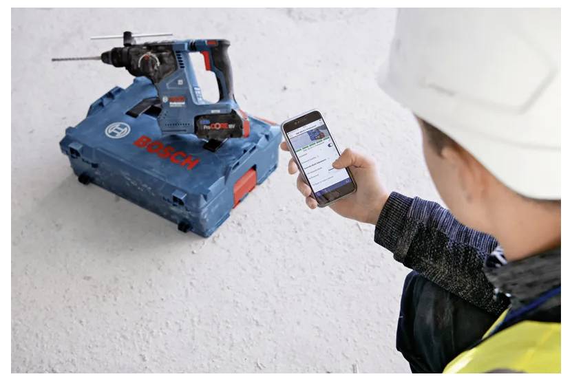 Ein Bauarbeiter benutzt eine Smartphone-App, um eine blaue Bosch-Akkuschrauber zu steuern, die auf ihrem Koffer auf einem Betonboden liegt.