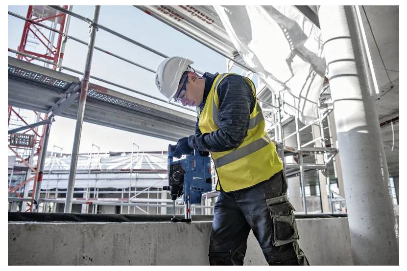 Ein Bauarbeiter in einer gelben Warnweste und einem Schutzhelm bedient einen Bohrer auf einer Betonoberfläche an einer Baustelle mit Gerüsten.