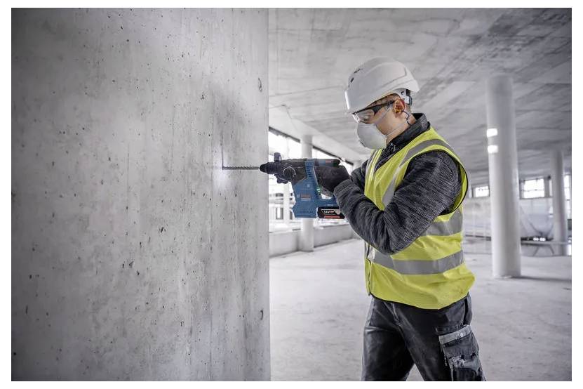 Ein Bauarbeiter mit Schutzhelm und Sicherheitsweste benutzt eine Bohrmaschine an einer Betonwand im Inneren eines unfertig gebauten Gebäudes.
