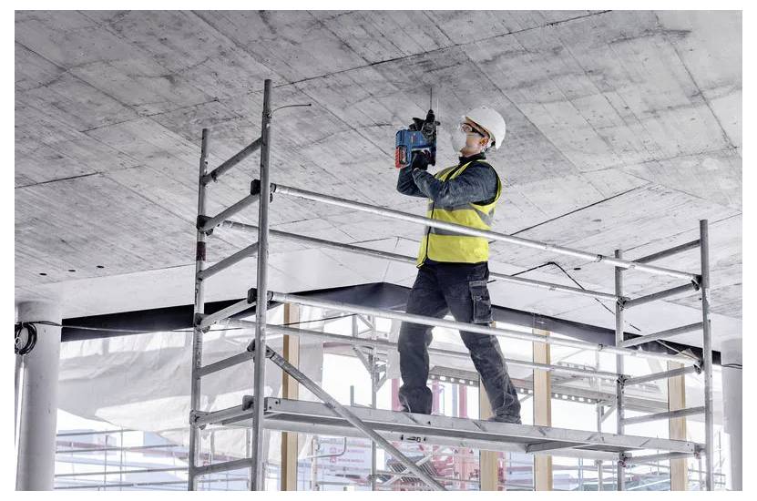 Ein Bauarbeiter mit Schutzhelm und Warnweste bedient ein Elektrowerkzeug an der Decke, während er auf einer Gerüstplattform auf einer Baustelle steht.