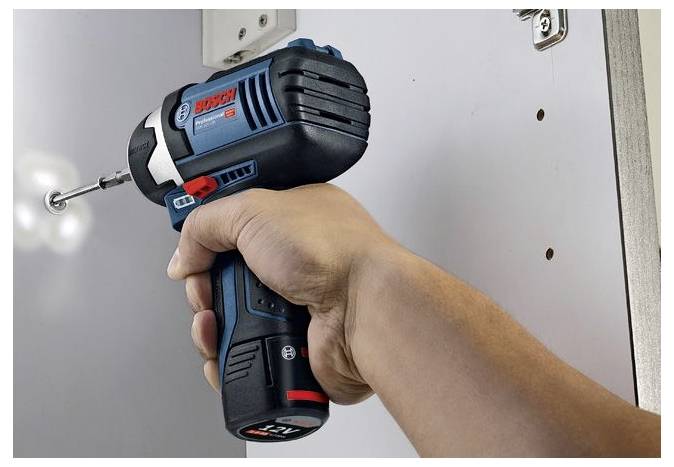 Eine Person verwendet eine blaue und schwarze kabellose Bohrmaschine, um in eine weiße Oberfläche zu schrauben. Die Bohrmaschine hat einen roten Knopf und eine Markenbeschriftung darauf.