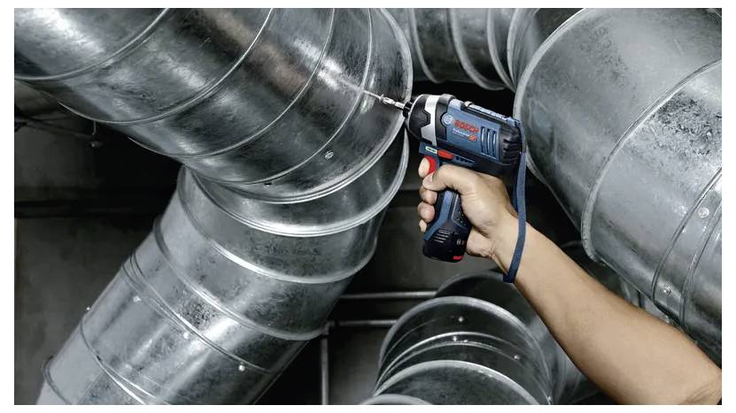 Eine Person verwendet einen kabellosen Akkuschrauber, um große Metallbelüftungskanäle zu befestigen und demonstriert damit die Leistungsfähigkeit des Werkzeugs in industriellen Umgebungen.