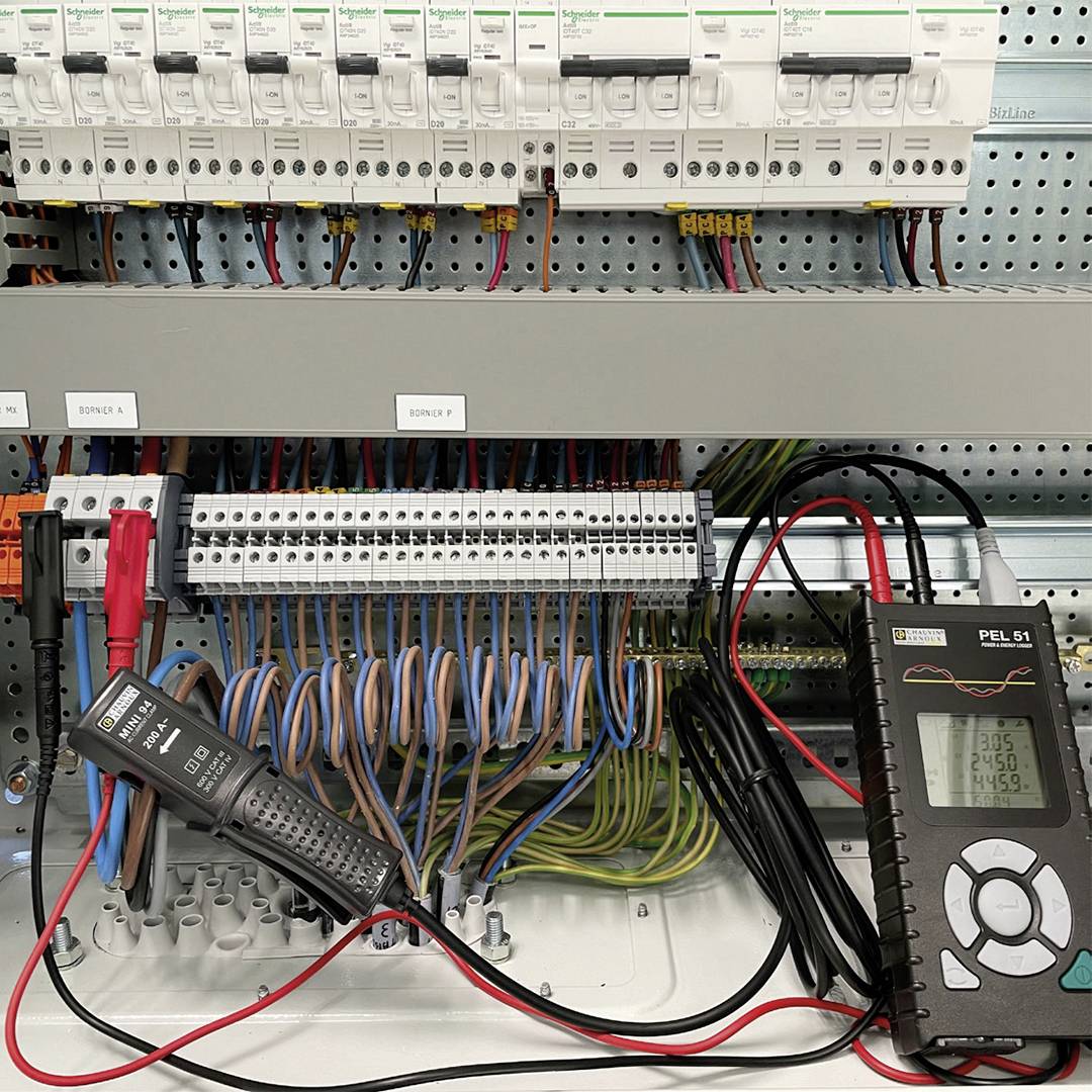 Ein offener Schaltschrank mit mehreren Sicherungen und Kabeln. Rechts ist ein digitales Messgerät, das den Energieverbrauch anzeigt.