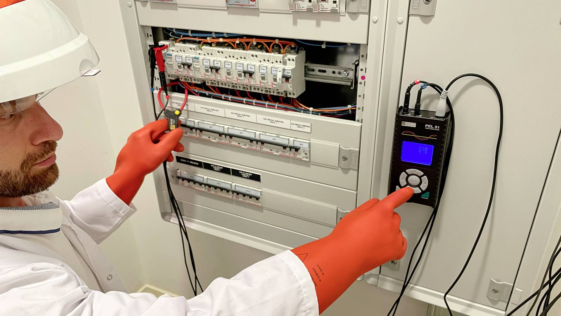 Ein Elektriker mit Schutzhelm und -handschuhen führt eine Messung an einem offenen Stromverteilerkasten mit einem Messgerät durch.