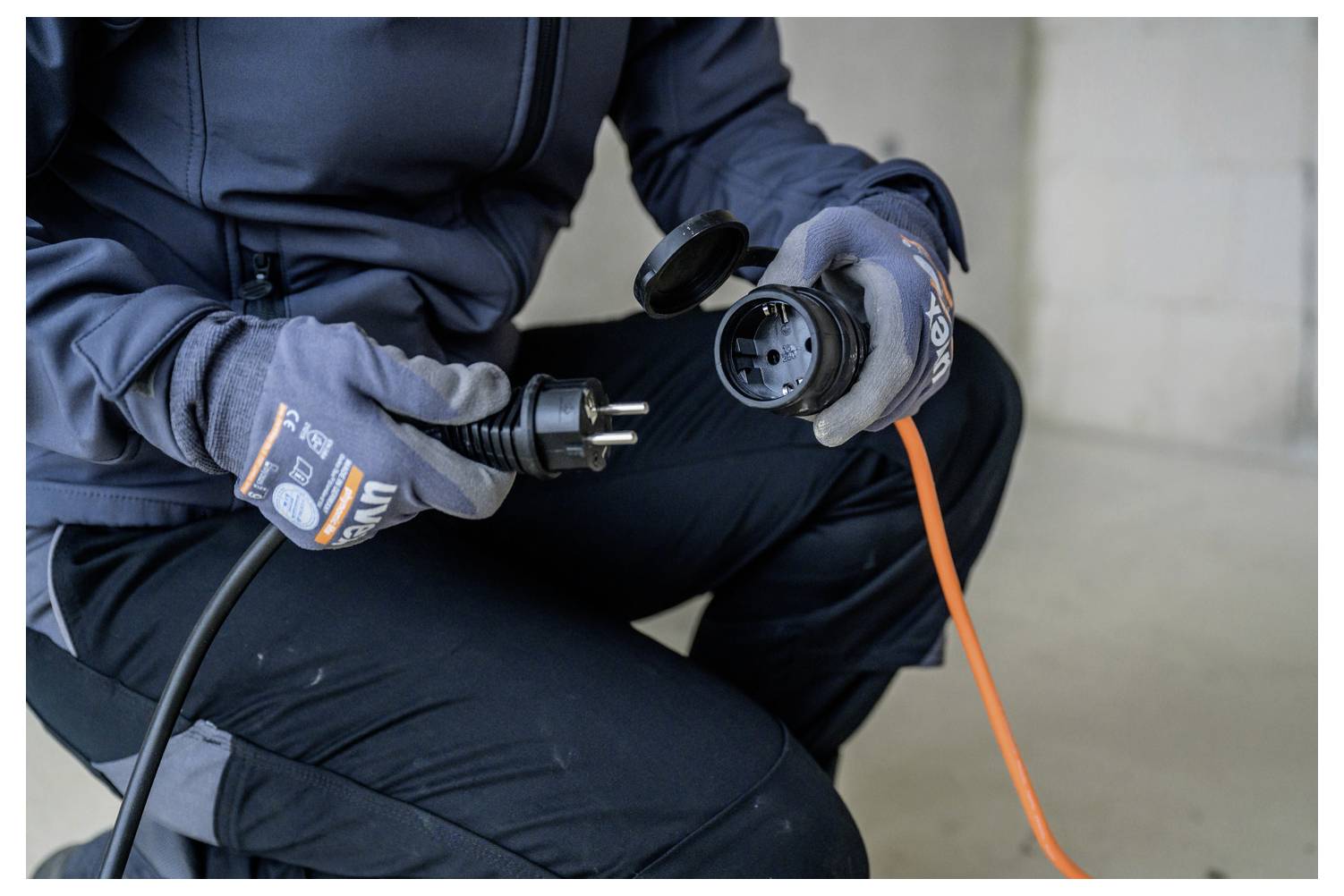 Eine Person mit Handschuhen steckt ein oranges Verlängerungskabel in einen schwarzen Stromstecker und bereitet eine elektrische Verbindung vor.