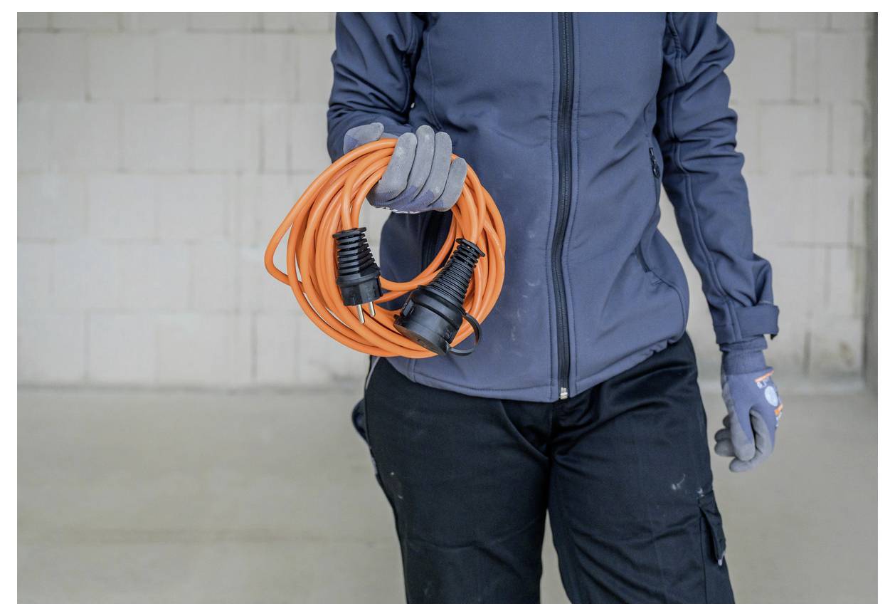 Person in Arbeitskleidung, die ein aufgerolltes orangefarbenes Verlängerungskabel mit zwei schwarzen Steckern gegen eine schlichte Innenwand hält.