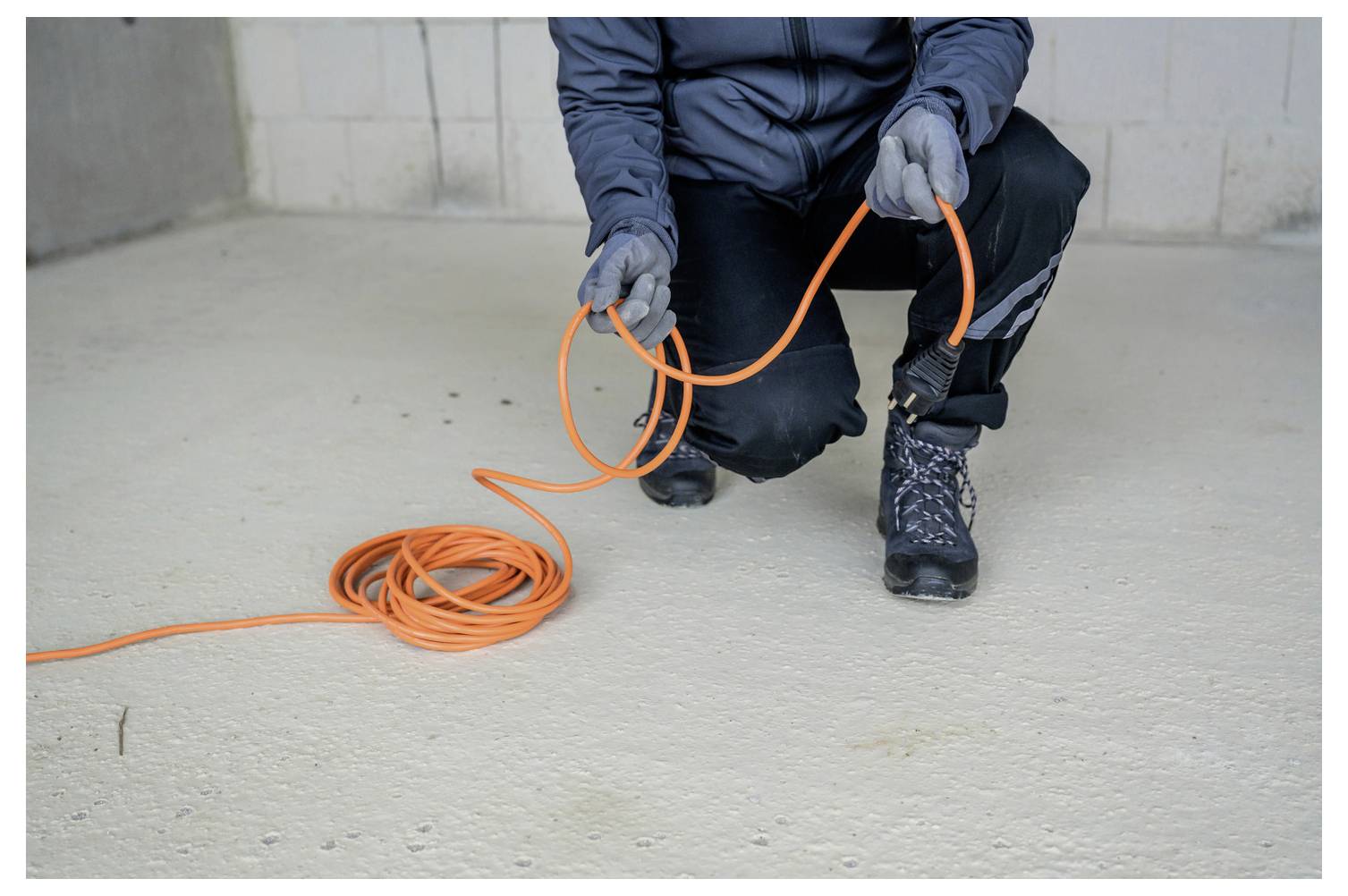 Arbeiter in Handschuhen kniet auf einem Betonboden, hält ein helles orange Kabel oder Verlängerungskabel und wickelt es ab.