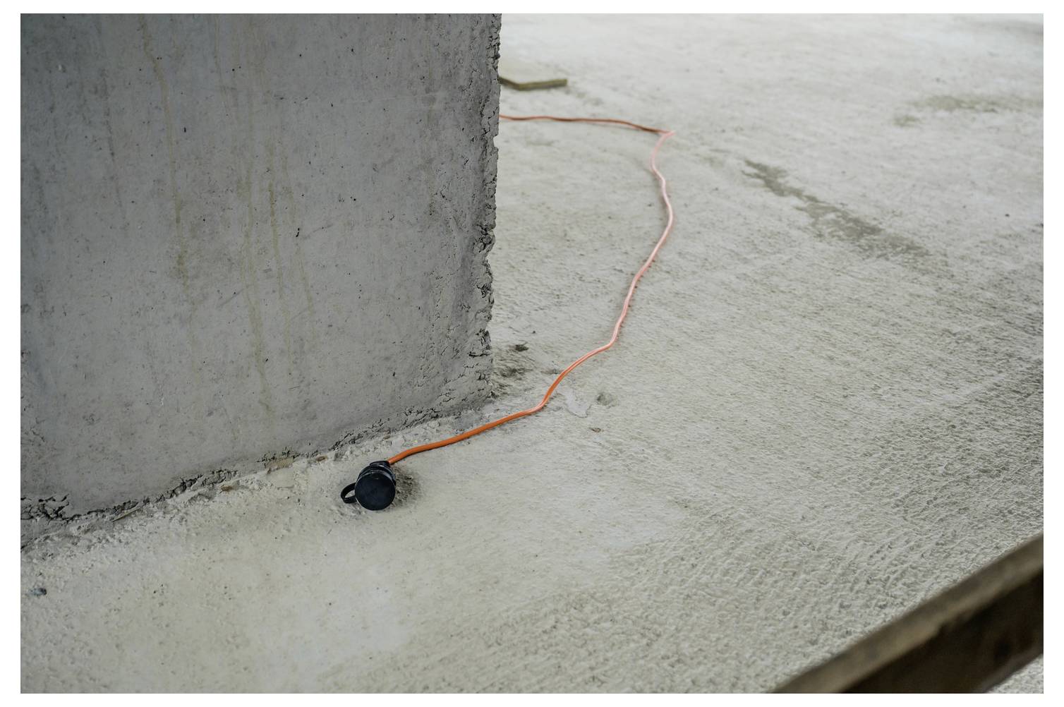 Oranges Verlängerungskabel mit schwarzem Stecker verläuft entlang eines Betonbodens neben einer großen Betonssäule.