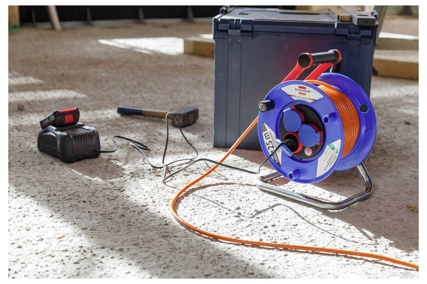 Tragbare Kabeltrommel mit orangem Kabel, Ladegerät und Werkzeugkasten auf einem Betonfußboden auf einer Baustelle.
