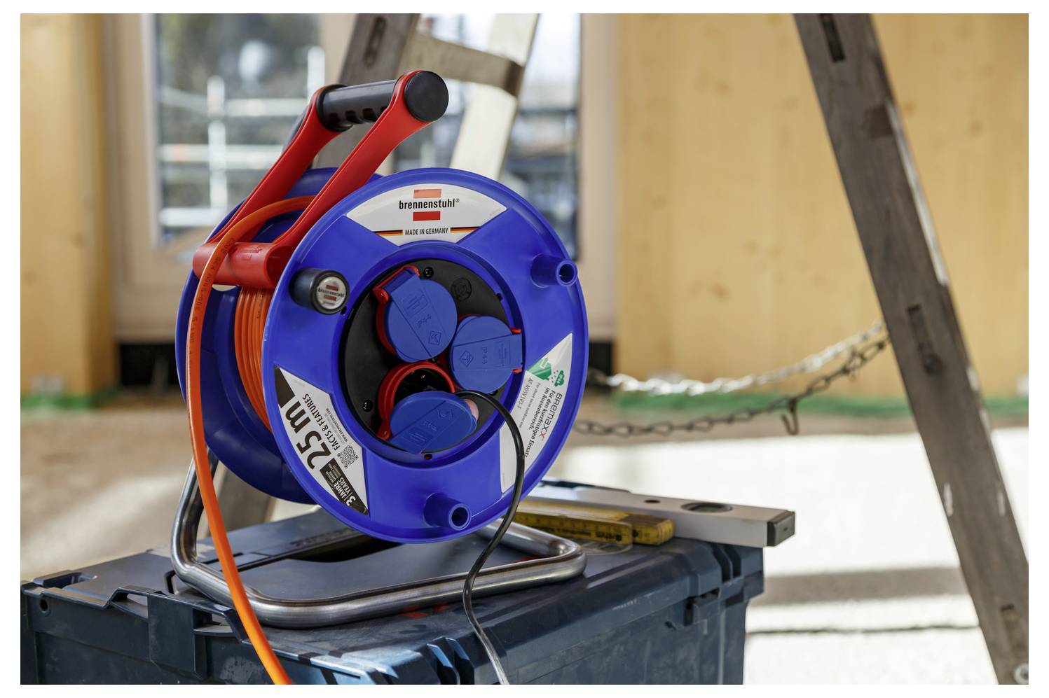 Blau-rote Kabelrolle auf einer Werkbank mit orangefarbenem Verlängerungskabel und Steckdosen in einer Werkstatt- oder Baustelle.