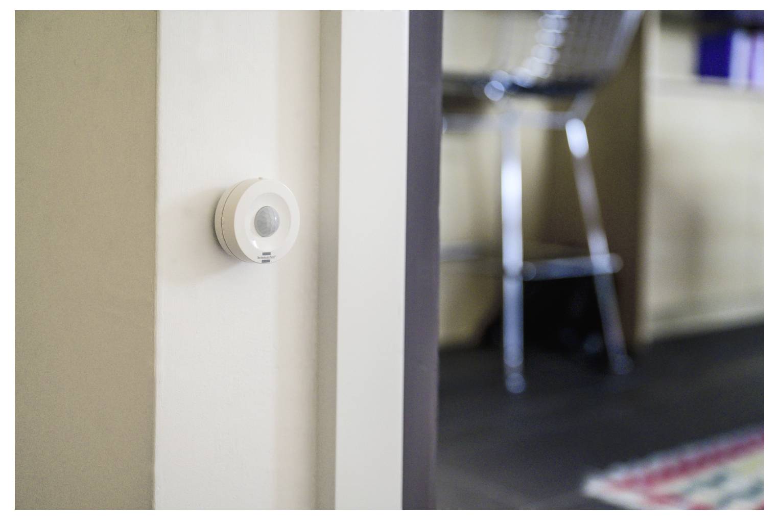 Kleiner weißer Bewegungsmelder an einer Wand neben einer Türöffnung montiert, mit einem unscharfen Zimmer und Stuhl im Hintergrund.