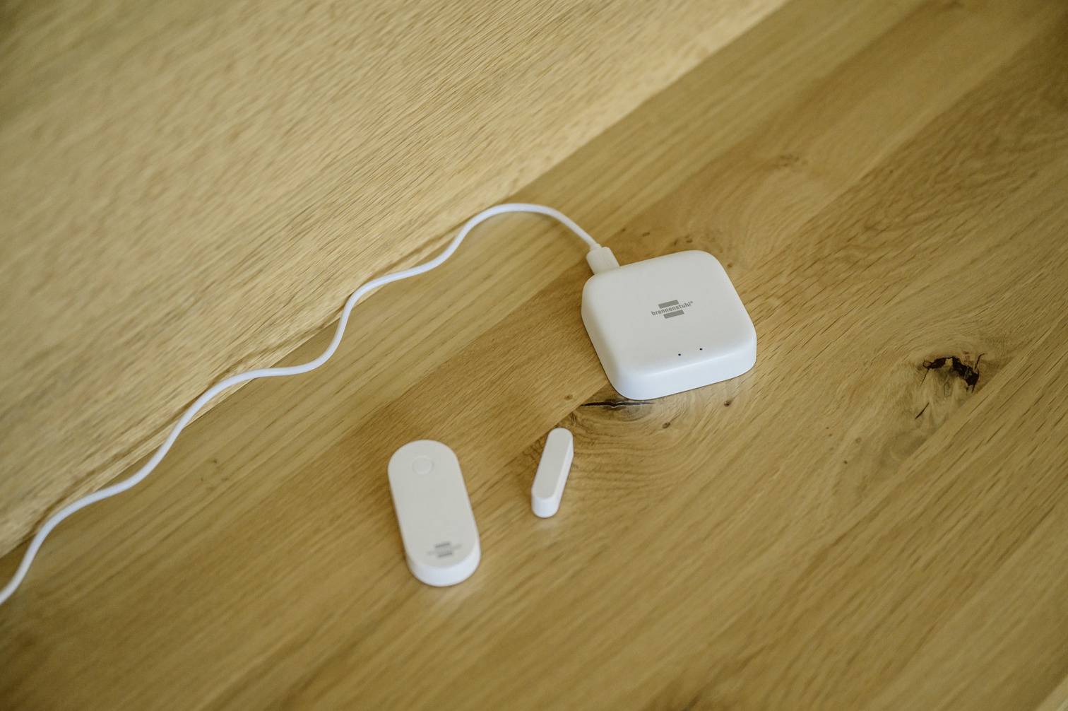Ein smartes Türsensor-Kit auf einem Holztisch zeigt den Hauptsensor, den Magnetkontakt und ein Hub-Gerät mit Kabelverbindung.