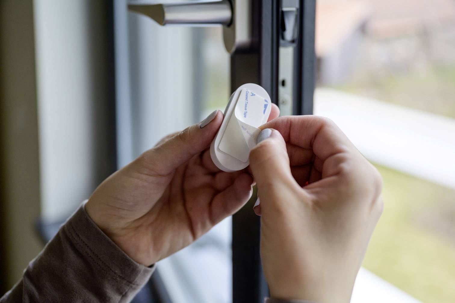 Eine Person installiert einen Sensor an einem Fensterrahmen. Der Sensor ist klein und weiß, und die Hände fixieren ihn vorsichtig an der Innenseite des Rahmens.