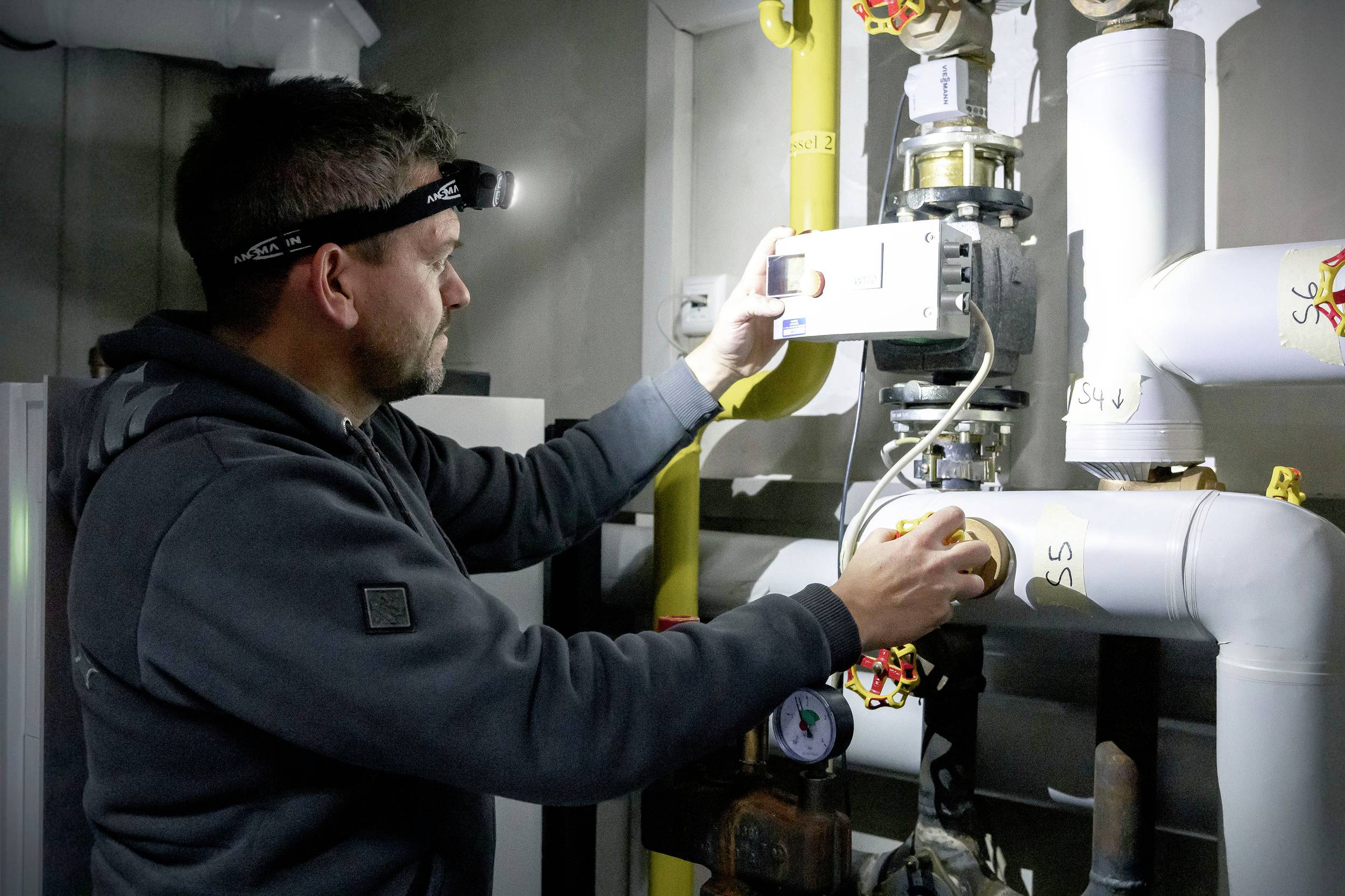 Ein Mann mit Stirnlampe überprüft ein Messgerät in einem technischen Raum, umgeben von Rohren und Heizungsanlagen.