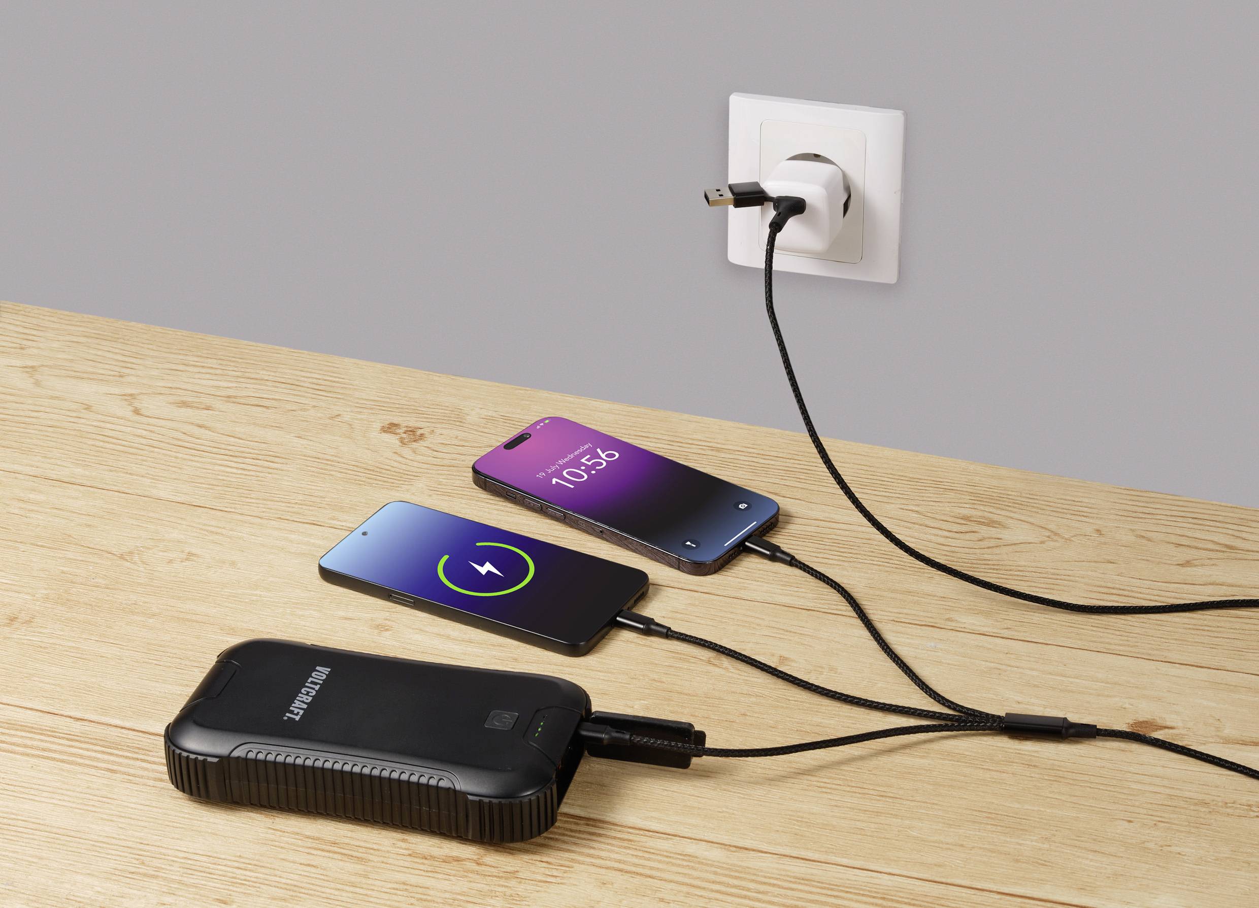 Zwei Smartphones liegen auf einem Holztisch und laden. Eines zeigt einen grünen Ladebildschirm, das andere die Uhrzeit 10:56 an. Sie sind mit einer Steckdose und einer Powerbank verbunden.