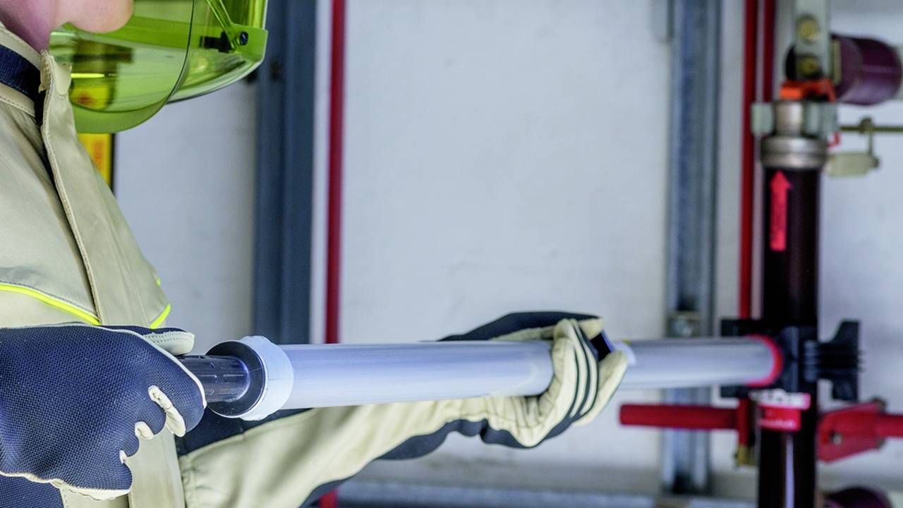 Person mit Schutzkleidung hält ein Rohr und arbeitet an einem Wasserleitungssystem in einem technischen Raum.