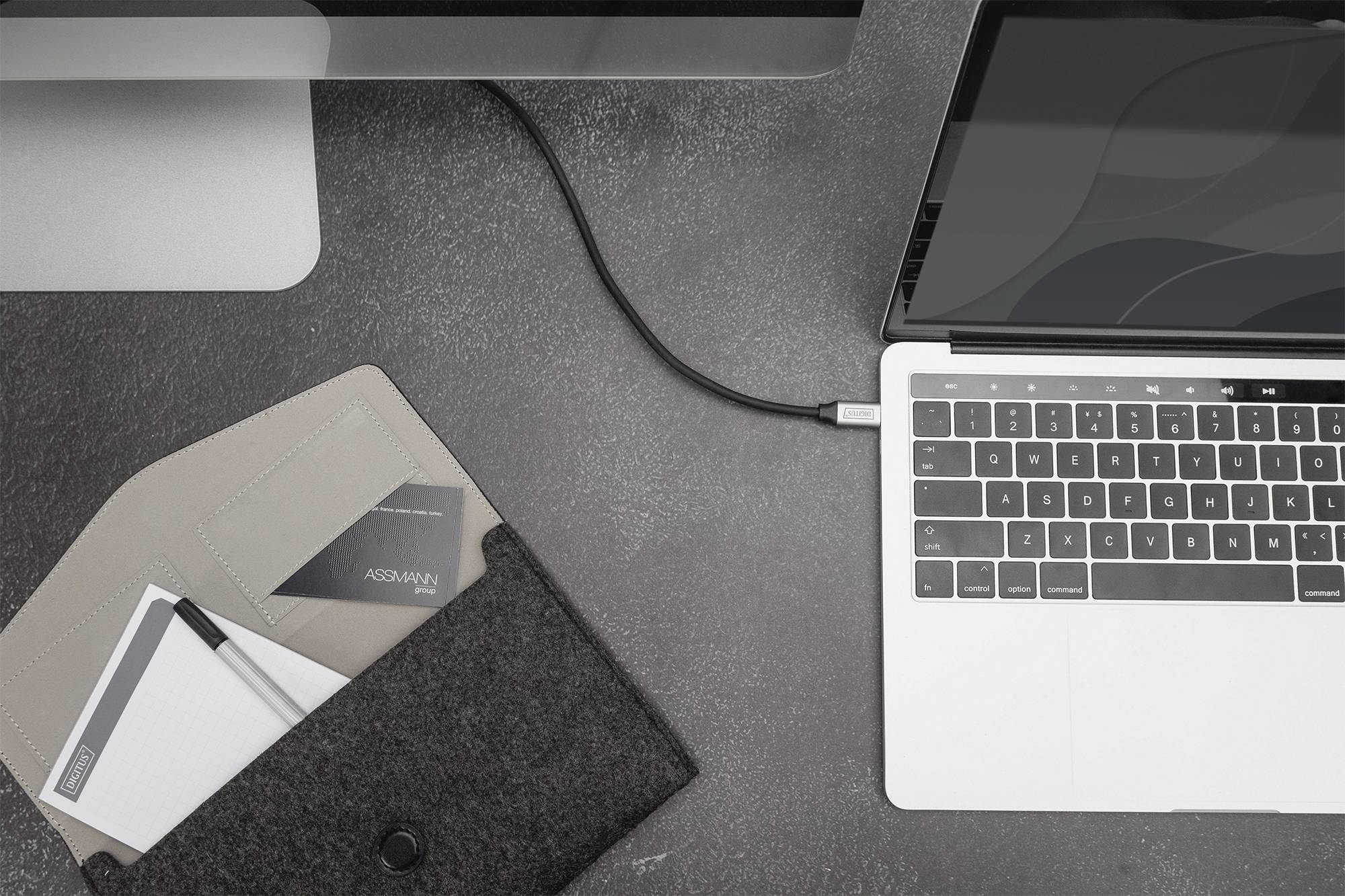 Laptop, der über ein Kabel an einen Desktop-Computer angeschlossen ist. Daneben eine Filztasche mit Notizblöcken auf grauem Tisch.