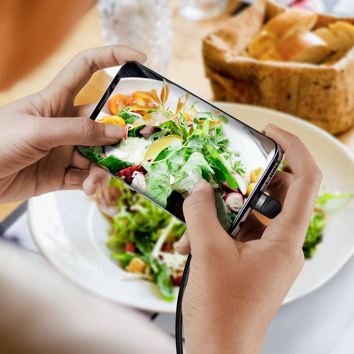Eine Person hält ein Smartphone und fotografiert ein buntes Salatgericht auf einem Teller. Im Hintergrund ist ein Glas und Brot zu sehen.