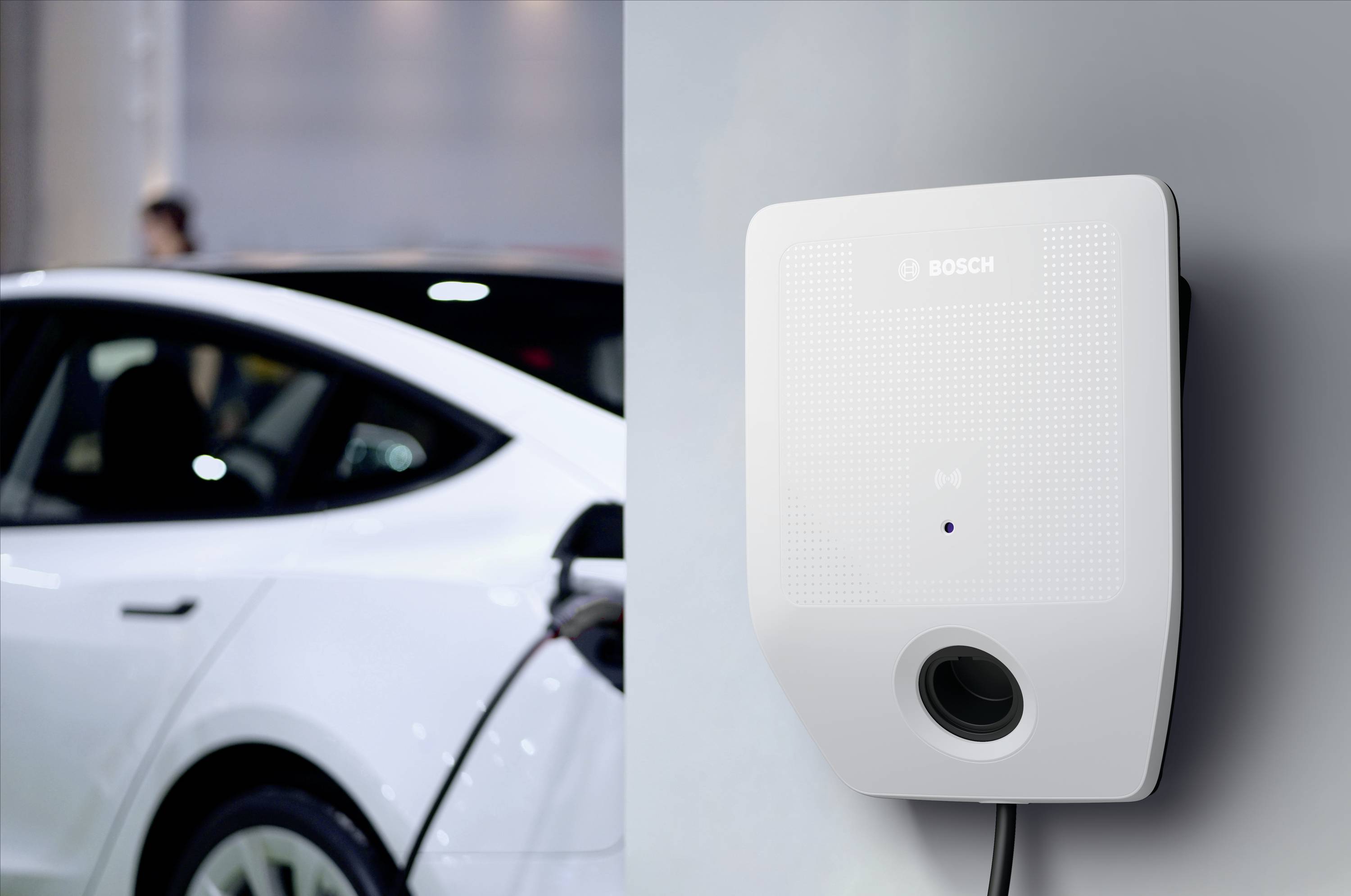 Ein E-Auto wird an einer weißen Ladestation geladen. Die Ladestation ist an einer Wand montiert, und das Auto ist teilweise sichtbar.