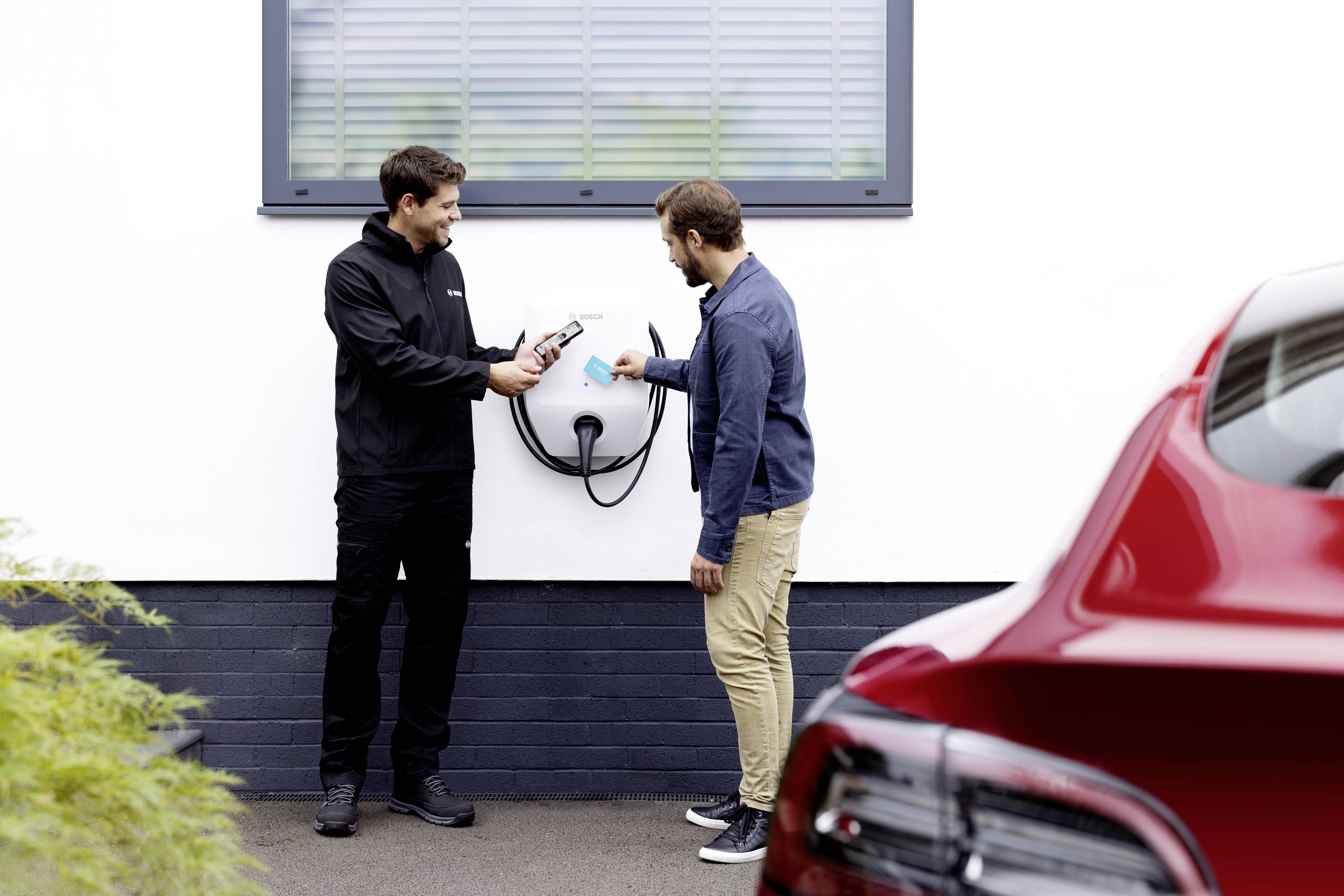 Ein Mann erklärt einem anderen die Verwendung einer Elektroauto-Ladestation an einer weißen Wand. Ein rotes Auto ist teilweise sichtbar.