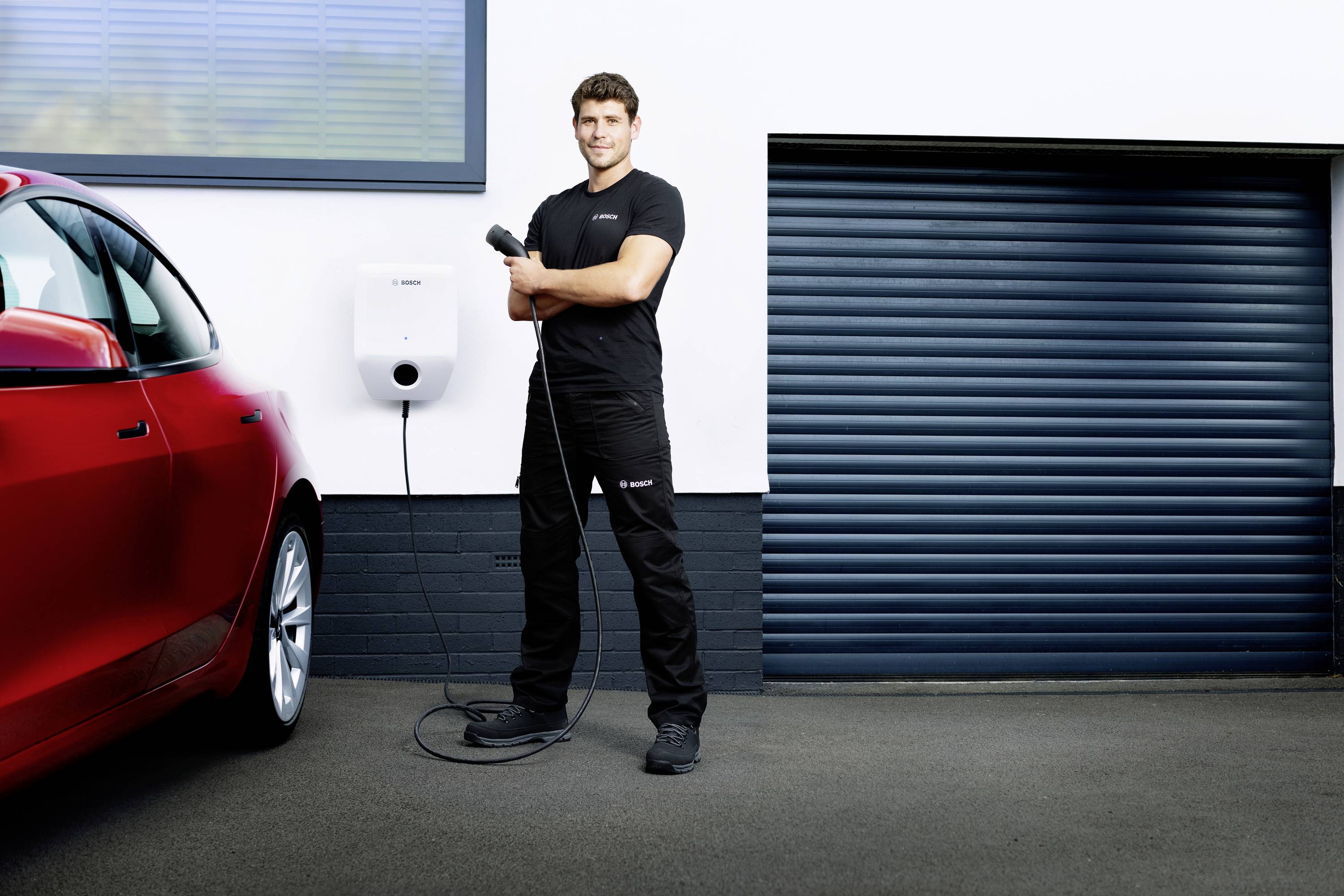 Ein Mann steht vor einer geschlossenen Garagentür, hält ein Ladekabel für ein E-Auto. Links ist ein rotes Auto teilweise sichtbar.