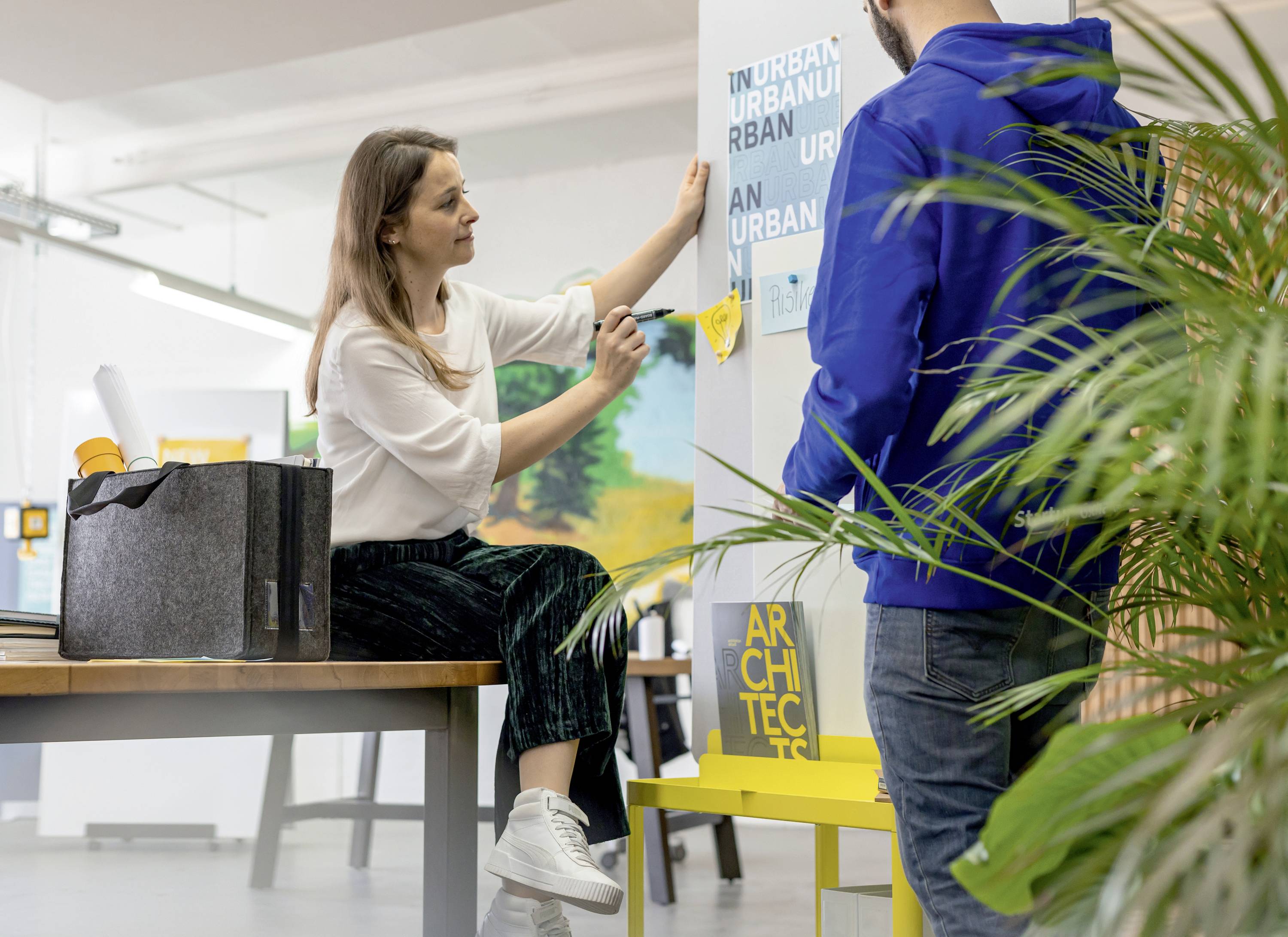Eine Frau sitzt an einem Tisch und ordnet mit einem Mann Poster auf einer Wand an. Sie diskutieren über Design oder Architektur.