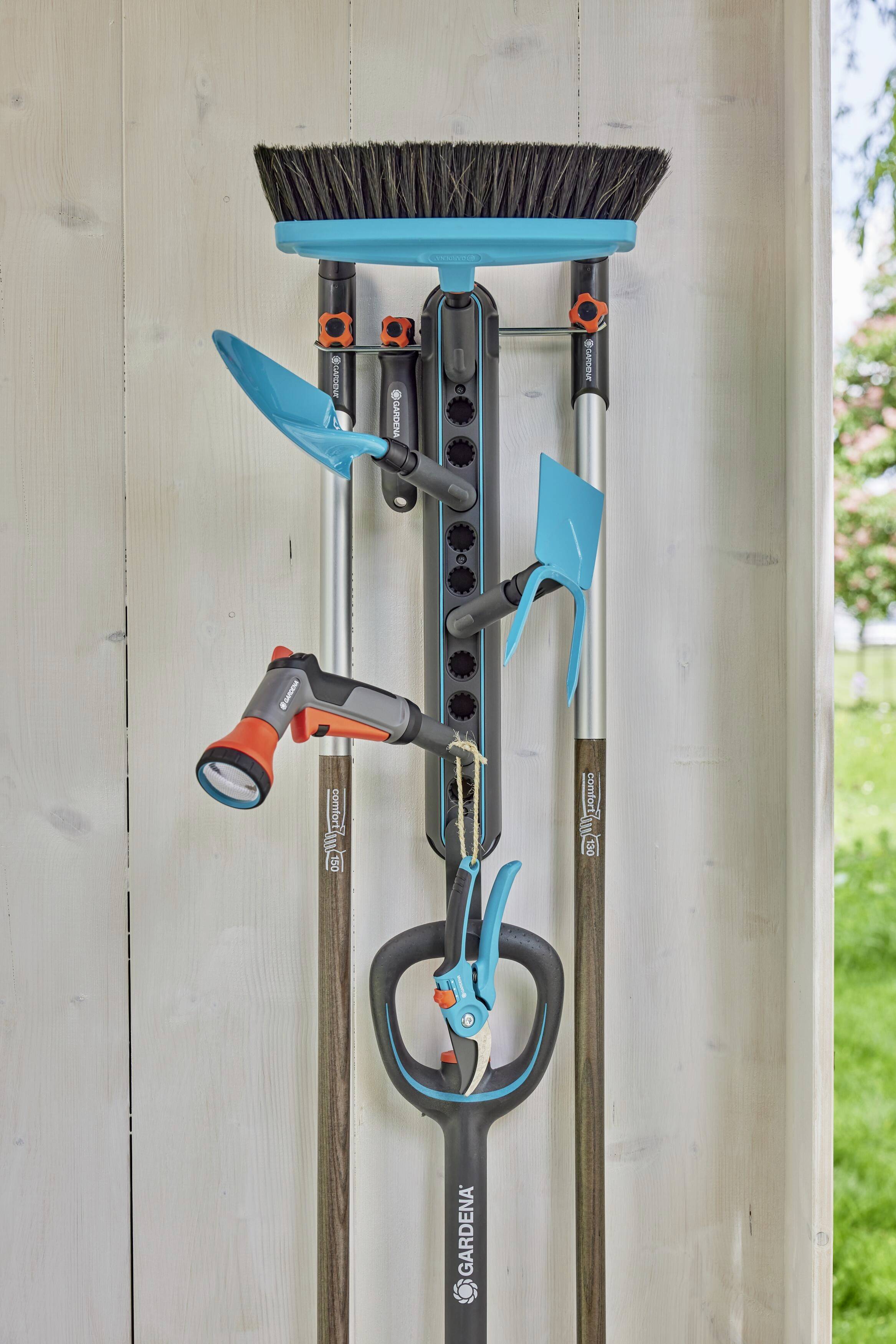 Ein Gartengerätehalter an einer Holzpaneelwand zeigt einen Besen, Schaufeln, eine Gartenschere und eine Gießspritze.