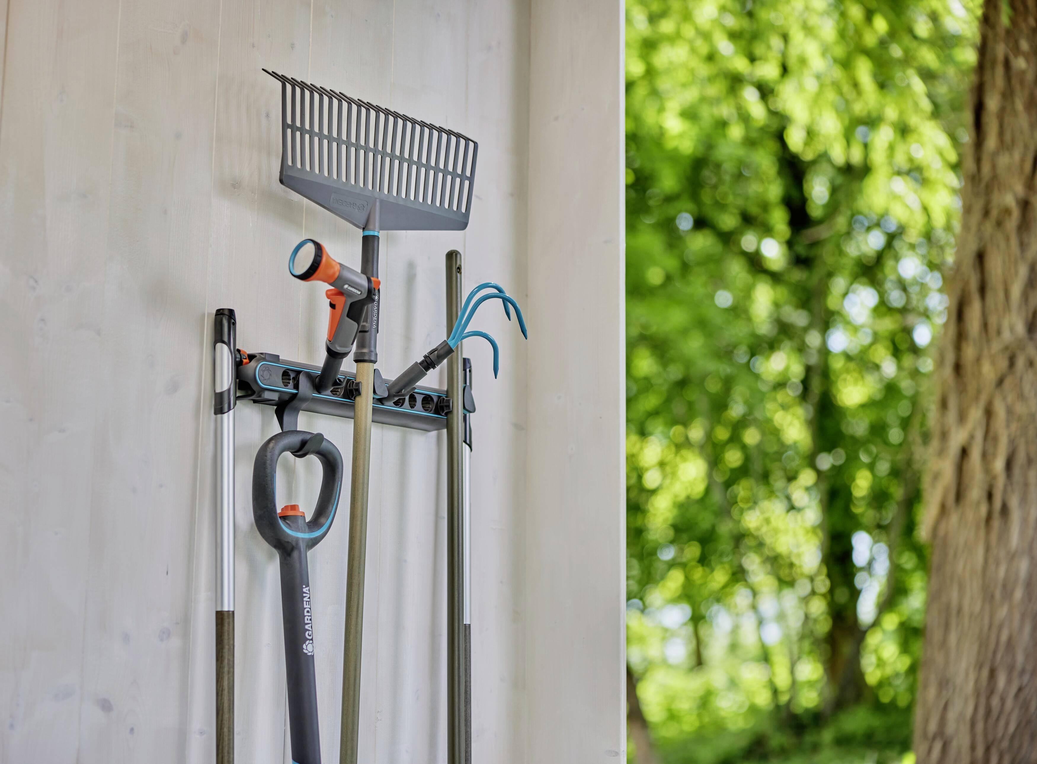 Gartenwerkzeuge mit Harke und Schlauch an weißer Wand montiert. Im Hintergrund unscharfes Grün eines Waldstücks.