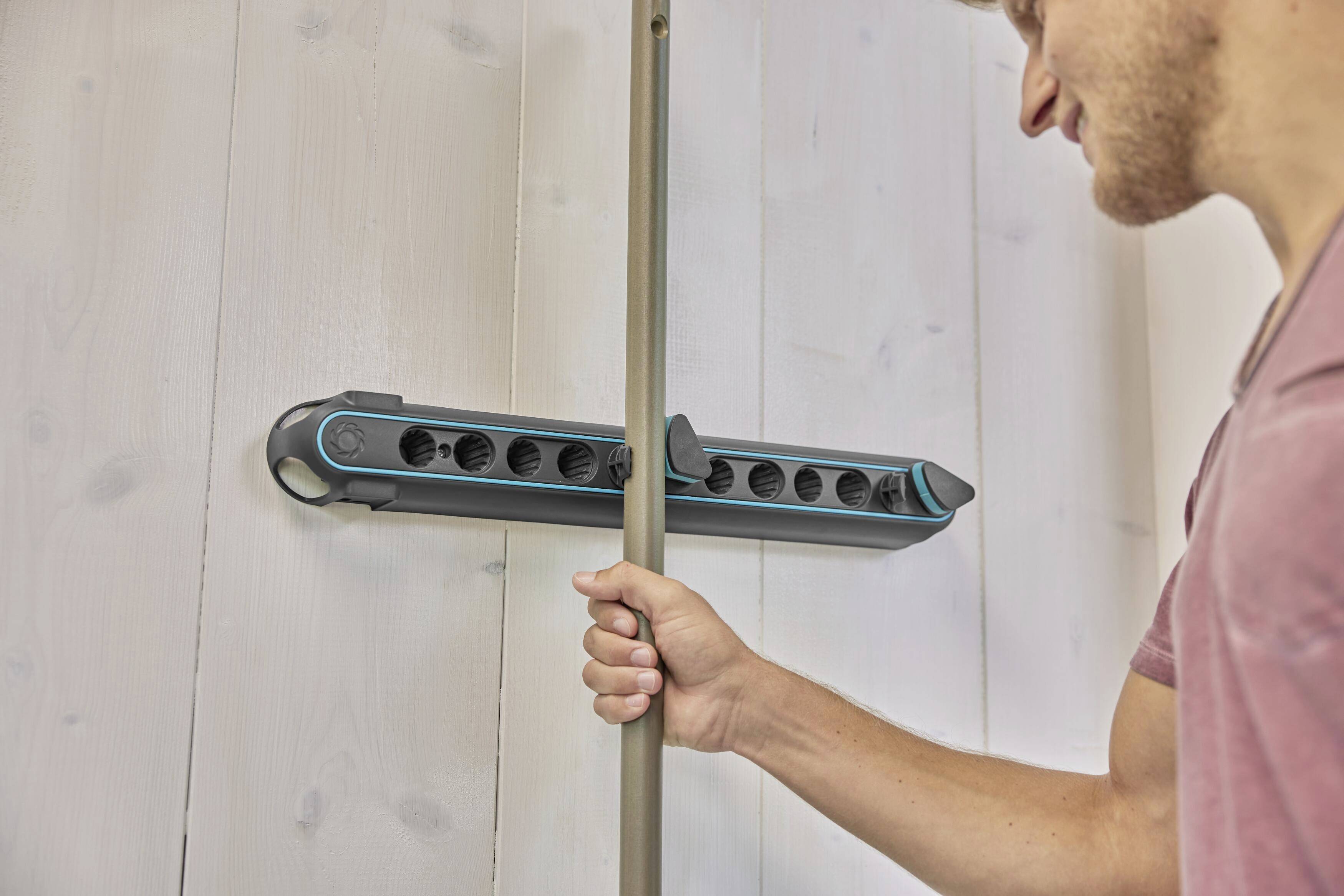 Eine Person montiert eine graue Stange an einer Wandhalterung mit mehreren Halterungen, vermutlich zur Aufbewahrung von Haushaltsgeräten.
