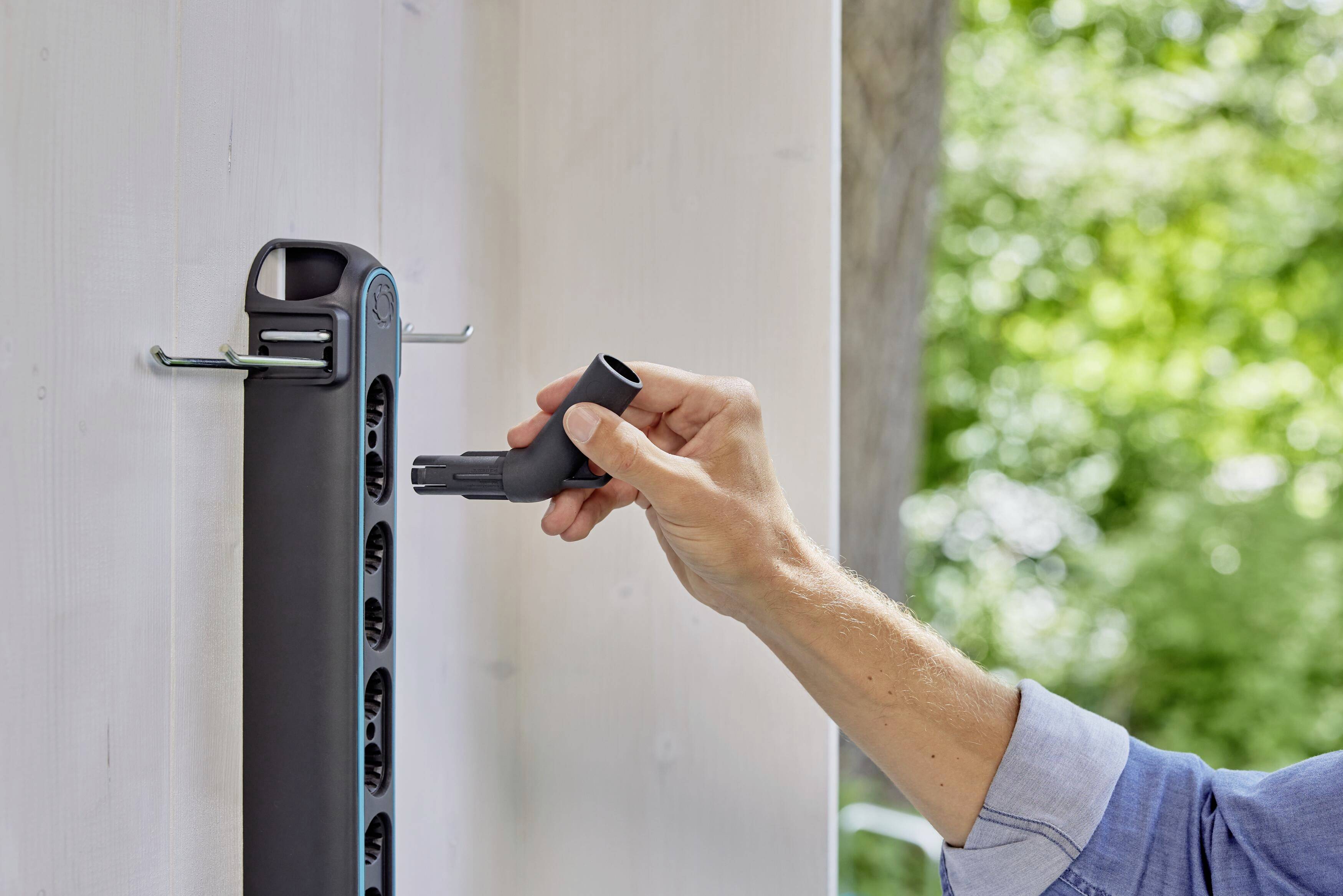 Eine Person hängt einen Gartenschlauch an einen Halter an einer weißen Wand auf, im Hintergrund ist grüne Vegetation sichtbar.