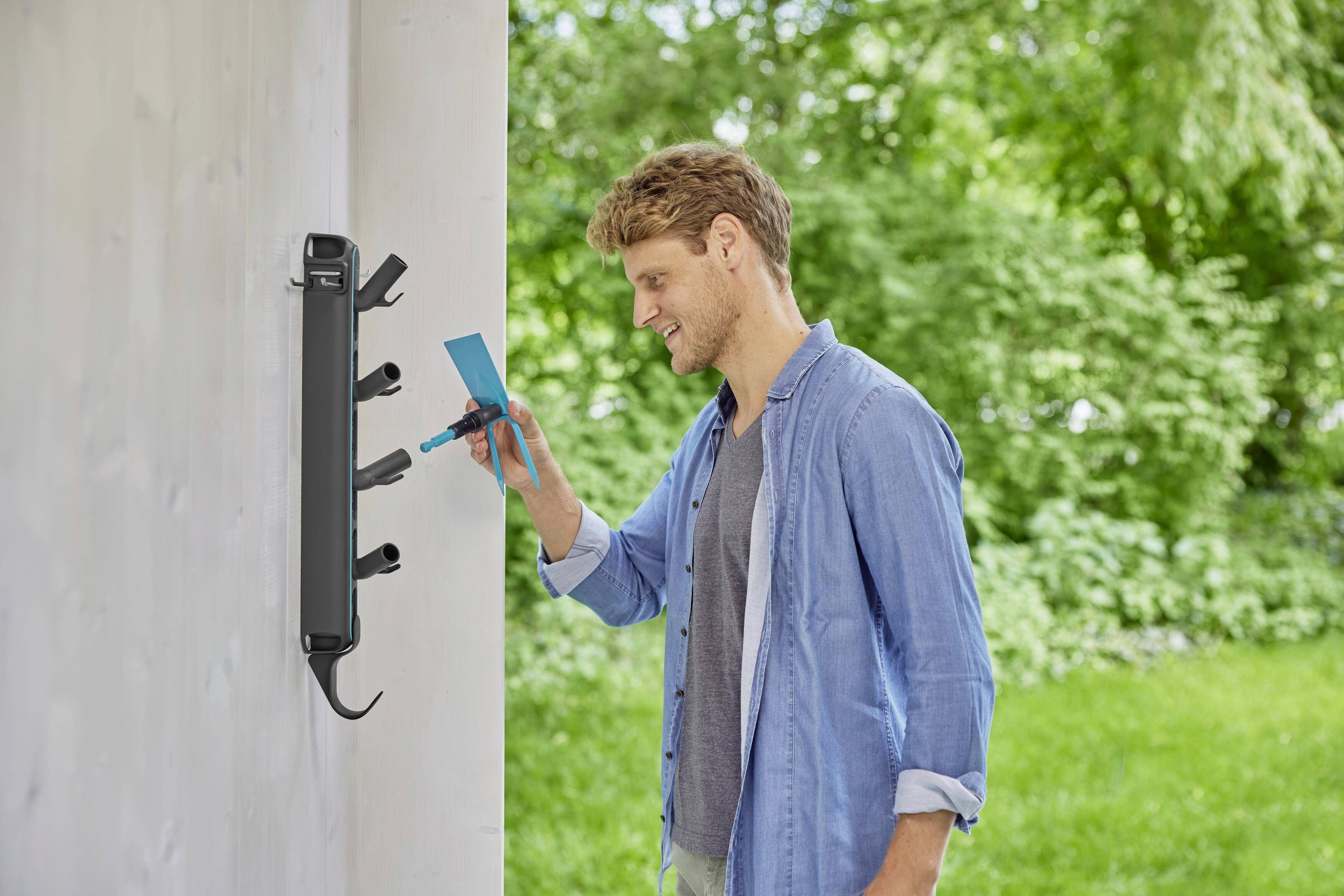 Ein Mann steckt ein Smartphone in eine Outdoor-Ladestation an einer Wand im Grünen. Lächelnd, hellblaues Hemd, sonniger Tag.