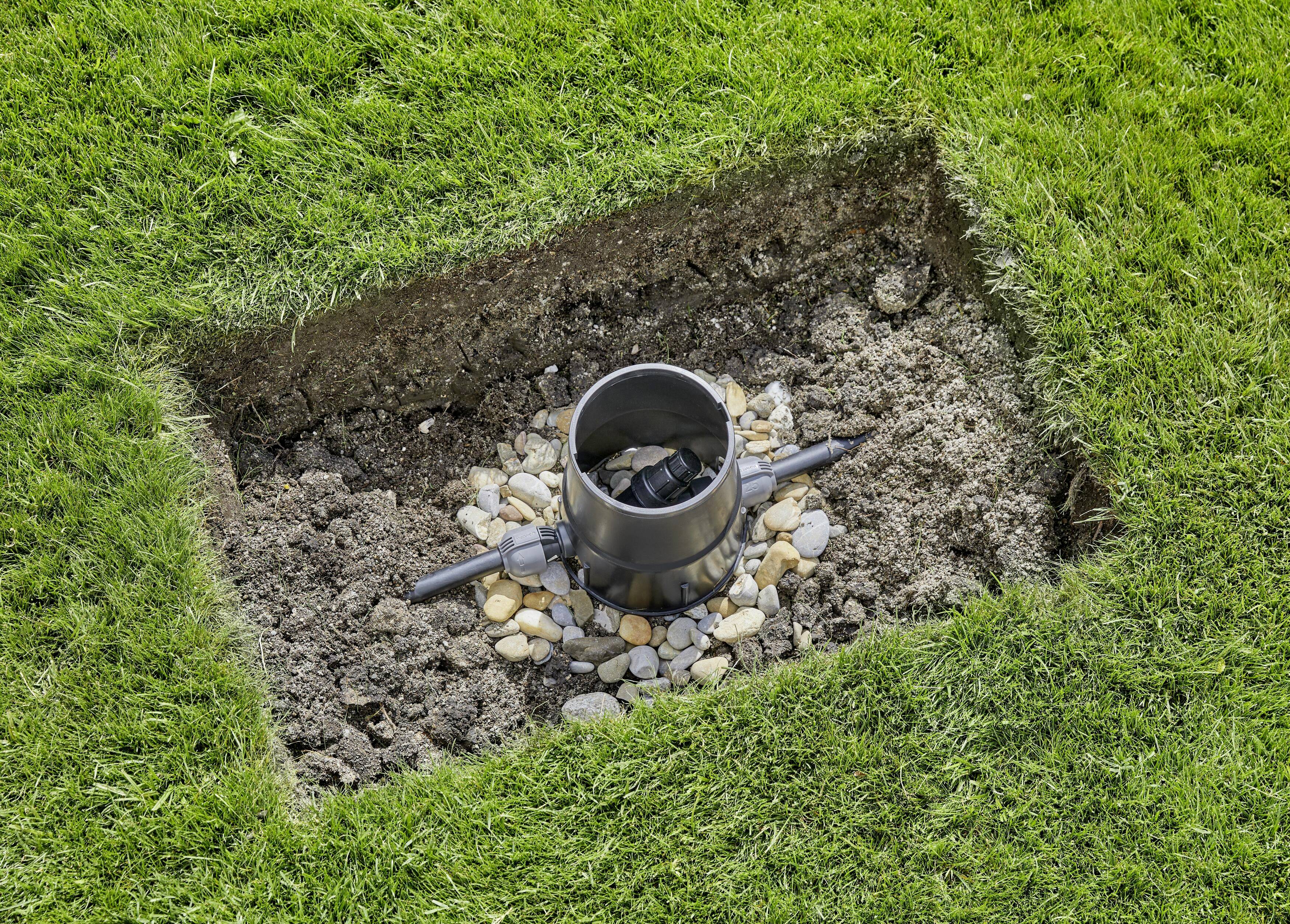 Ein quadratisches Loch im Rasen zeigt ein unterirdisches Sprinklersystem. Im Zentrum ist ein rundes Ventil, umgeben von Kieselsteinen.