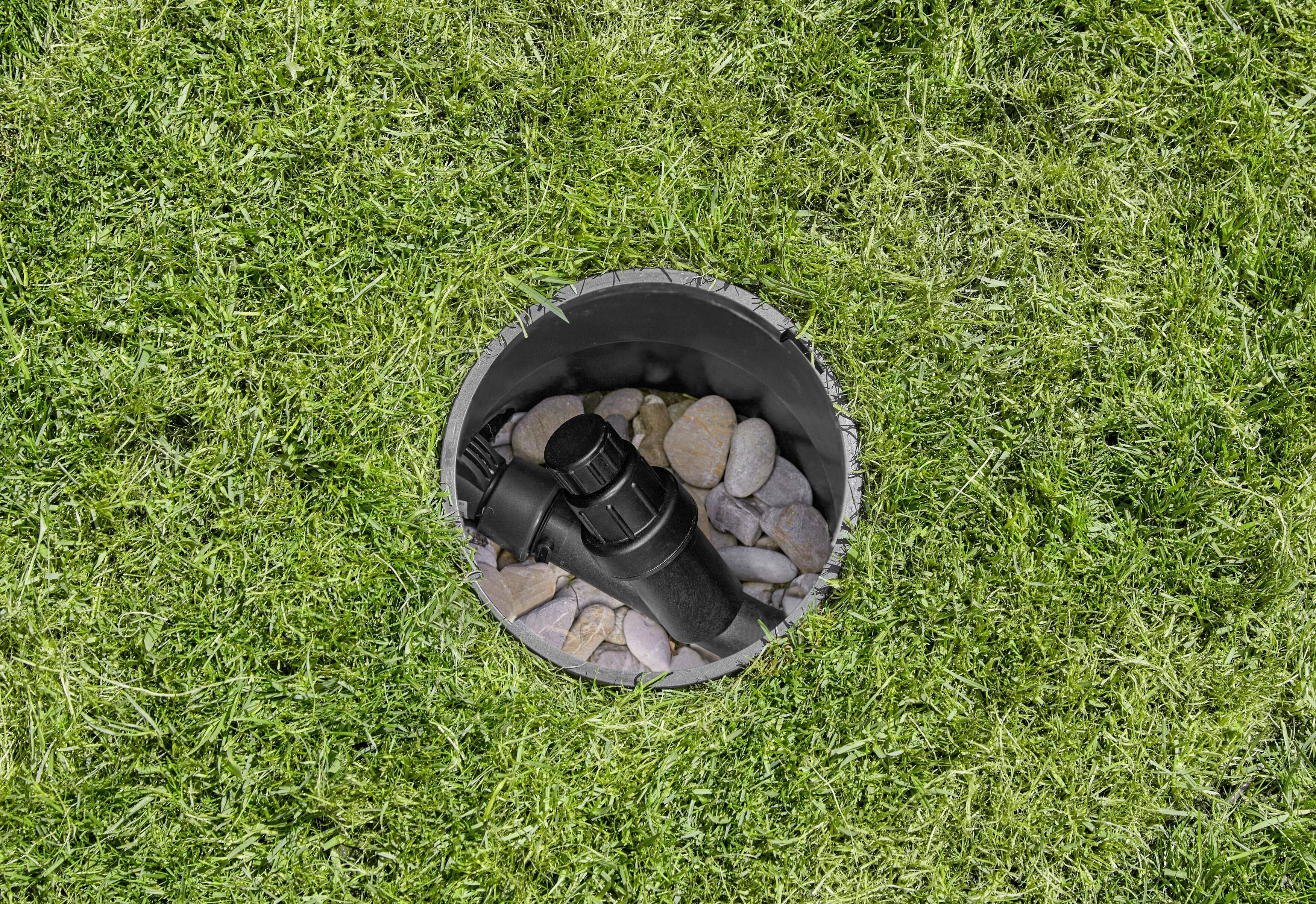 Ein schwarzer Sprinklerkopf ist von einer runden Öffnung auf einer Rasenfläche umgeben, mit Steinen im Inneren der Öffnung.