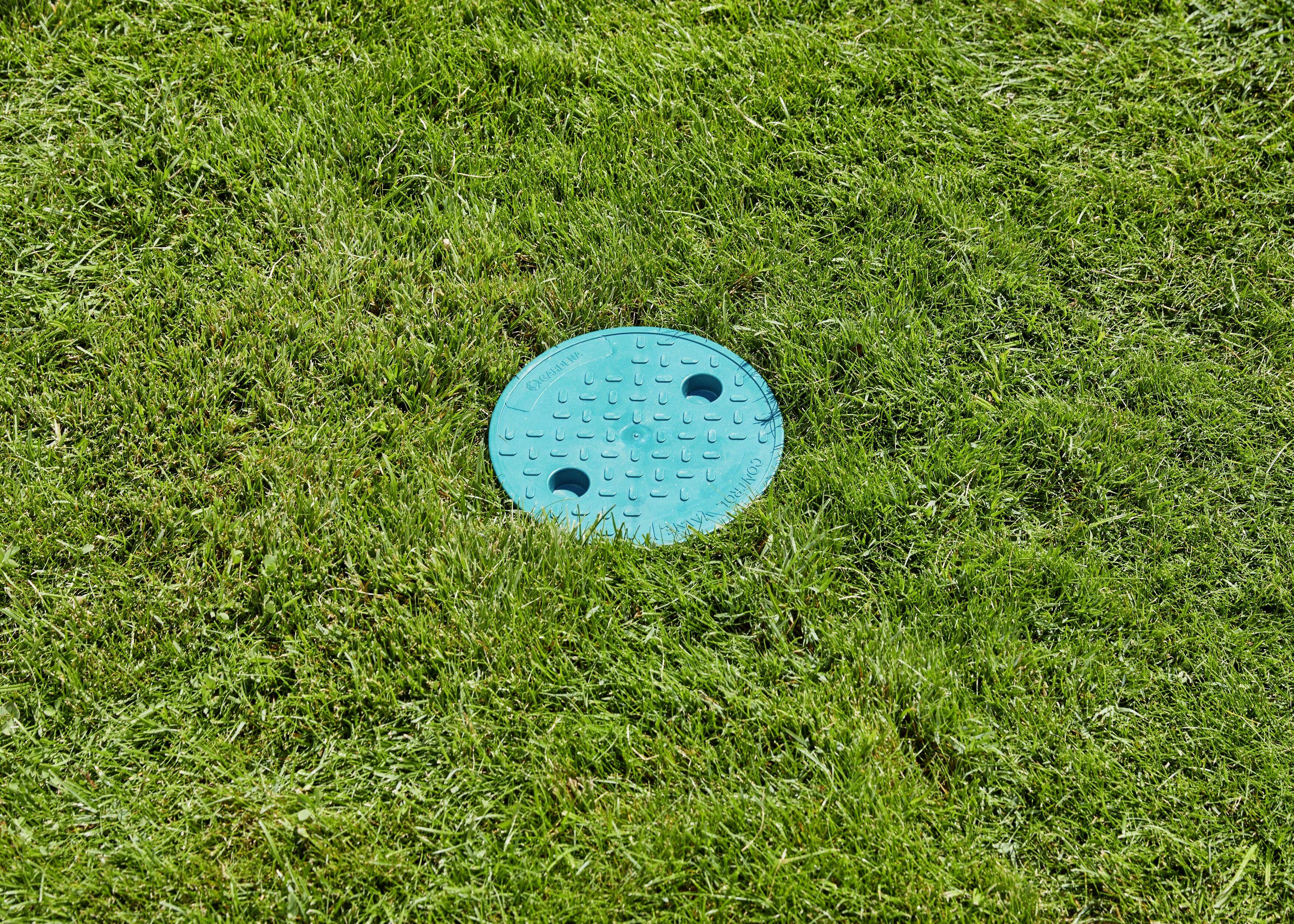 Ein blauer Abwasserkanaldeckel auf einem grünen Rasen. Der Deckel hat zwei Löcher und ist von geschnittenem Gras umgeben.