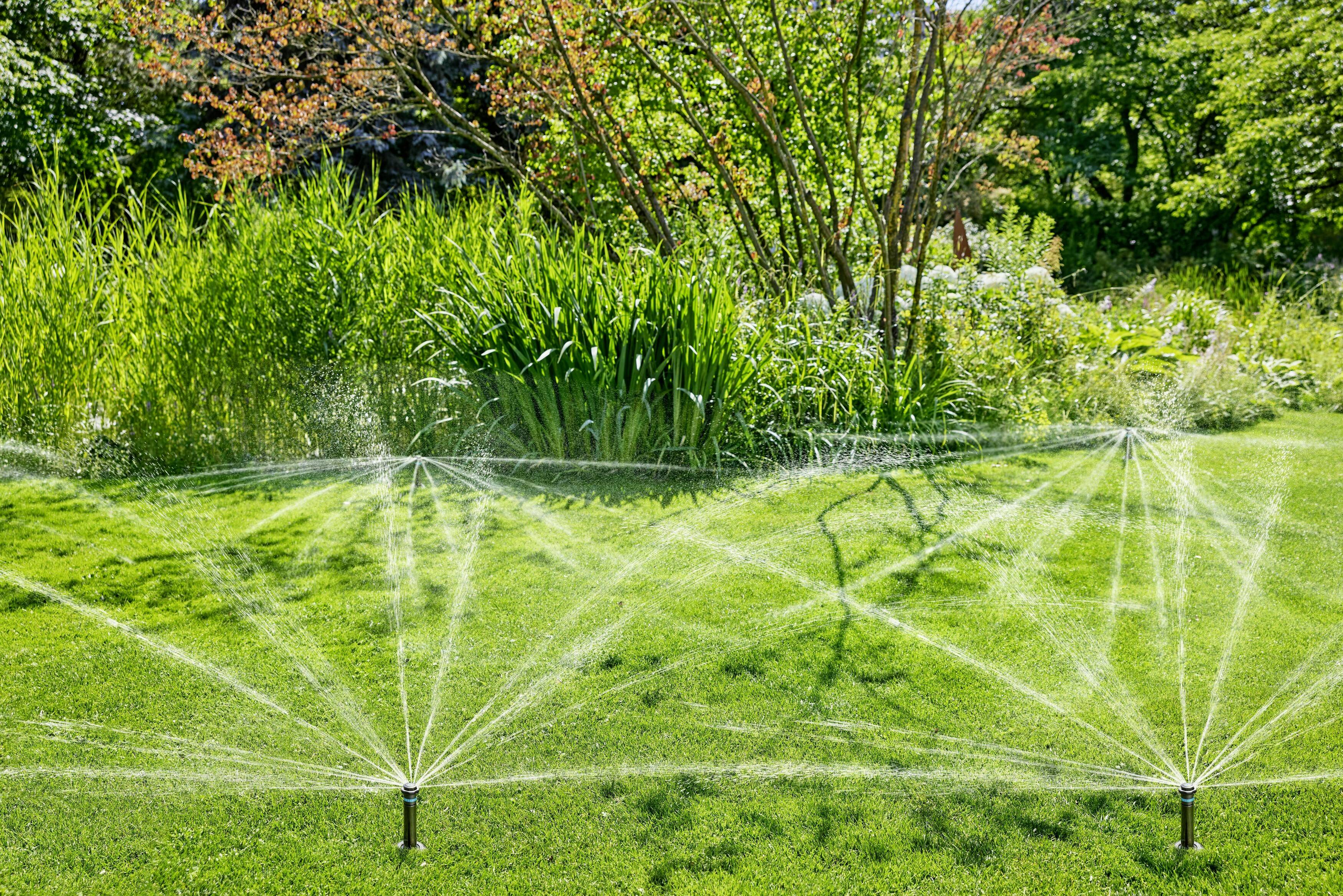 Ein Garten mit grünem Gras wird von zwei laufenden Sprinklern bewässert, umgeben von Bäumen und Büschen im Hintergrund.