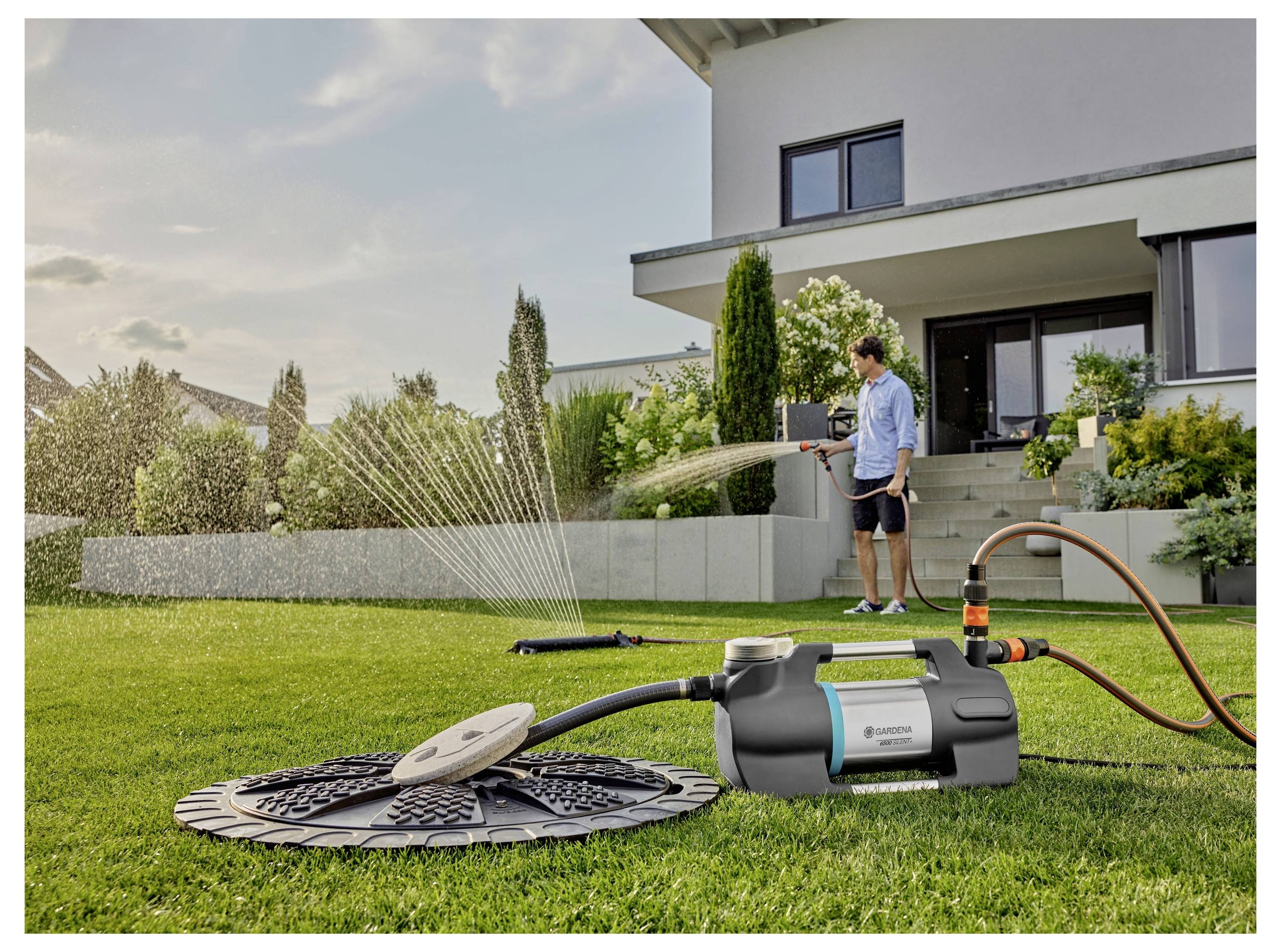 Eine Person bewässert einen Rasen mit einem Schlauch in einer modernen Gartenanlage. Ein Bewässerungssystem auf dem Gras verteilt Wasser gleichmäßig über die Fläche.
