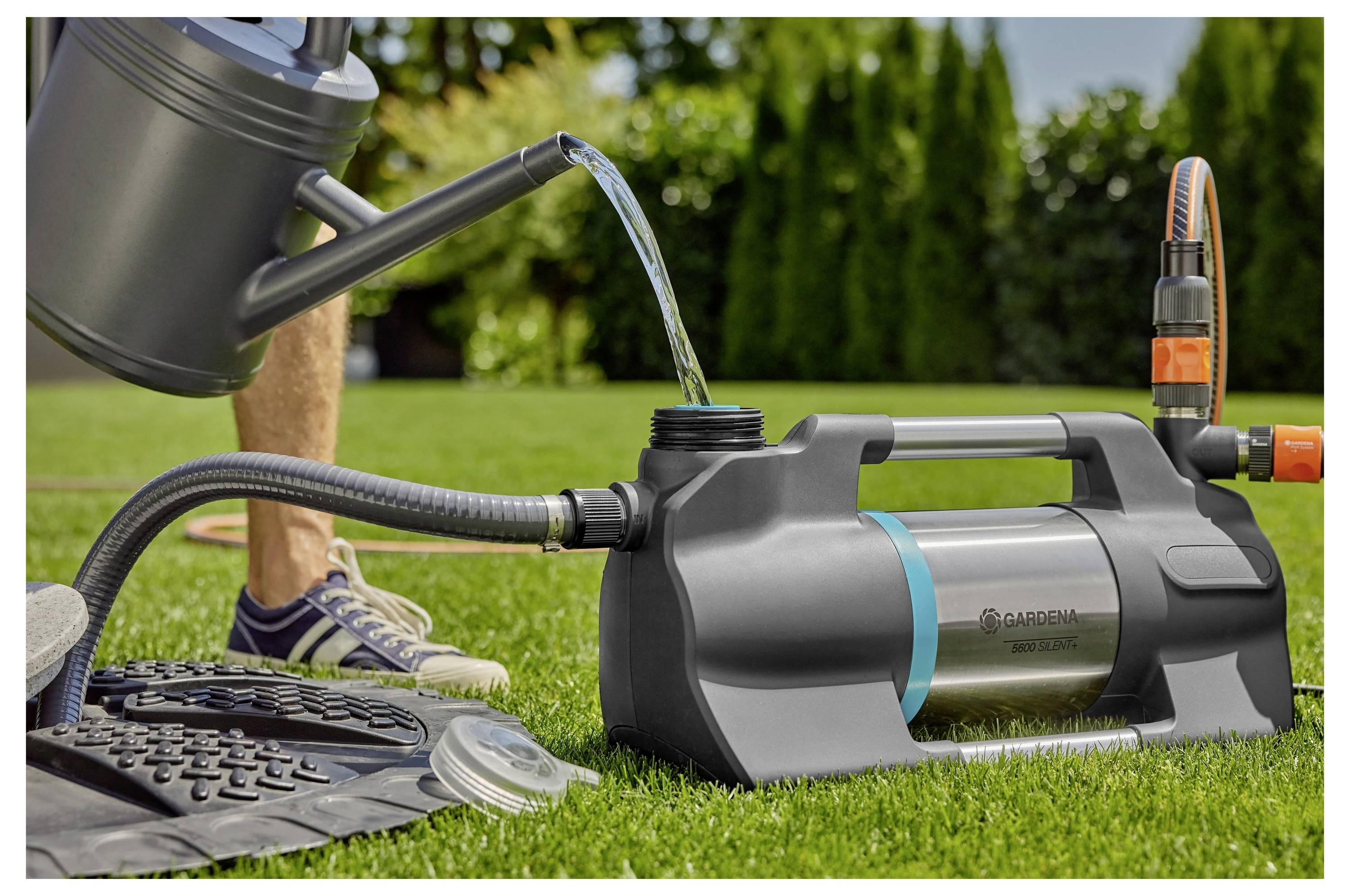 „Eine Gartenpumpe, die Wasser aus einem Reservoir in eine Gießkanne auf einem Rasen umleitet, mit einer Person im Hintergrund, die Turnschuhe trägt.
