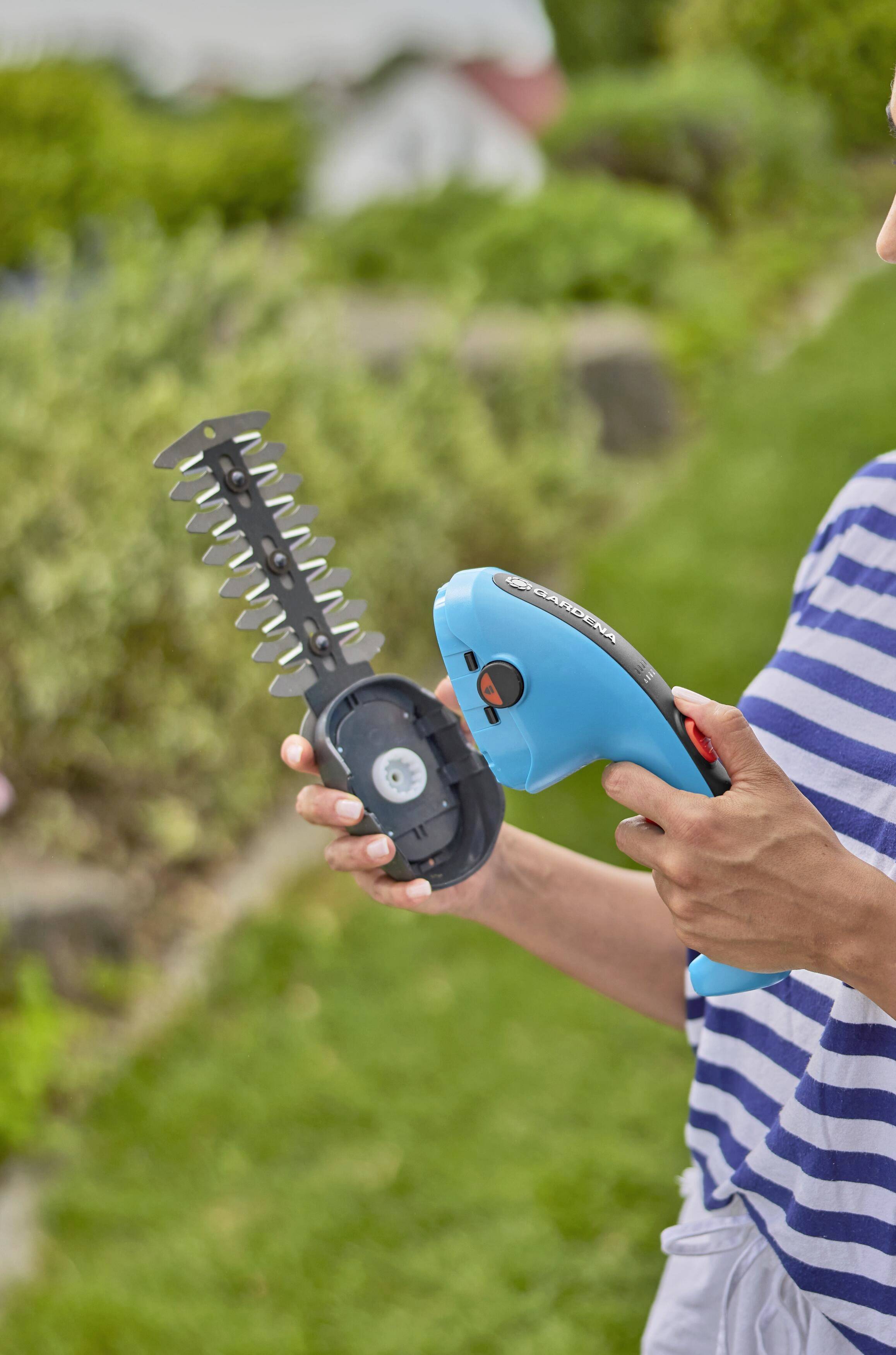 Person hält einen blauen, akkubetriebenen Heckenschneider; im Hintergrund unscharfe Gartenvegetation.
