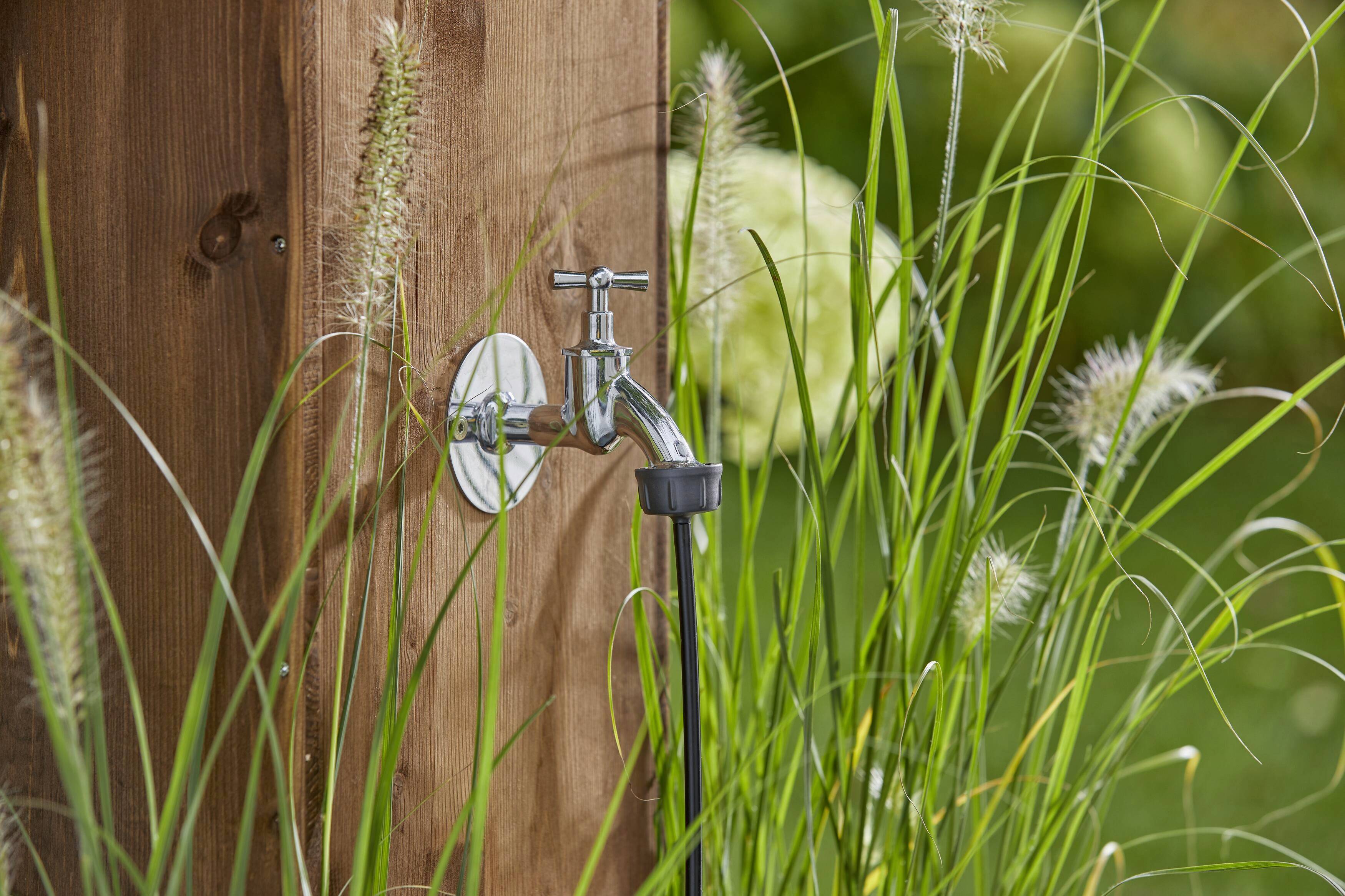 Ein Wasserhahn ist an einem Holzpfosten montiert, umgeben von hohem Gras. Der Hintergrund zeigt eine grüne, naturbelassene Umgebung.